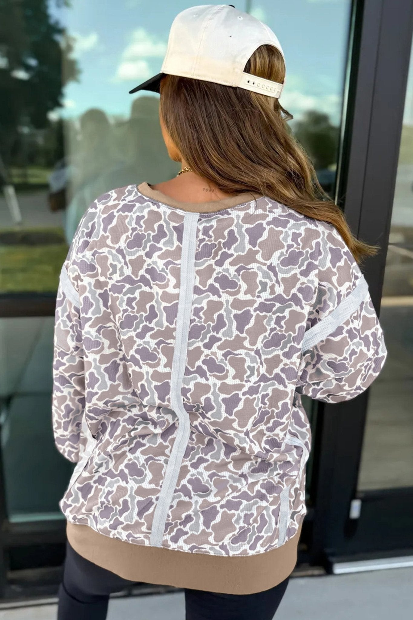 Back view of woman in taupe-white abstract zip jacket, black leggings, and white cap.