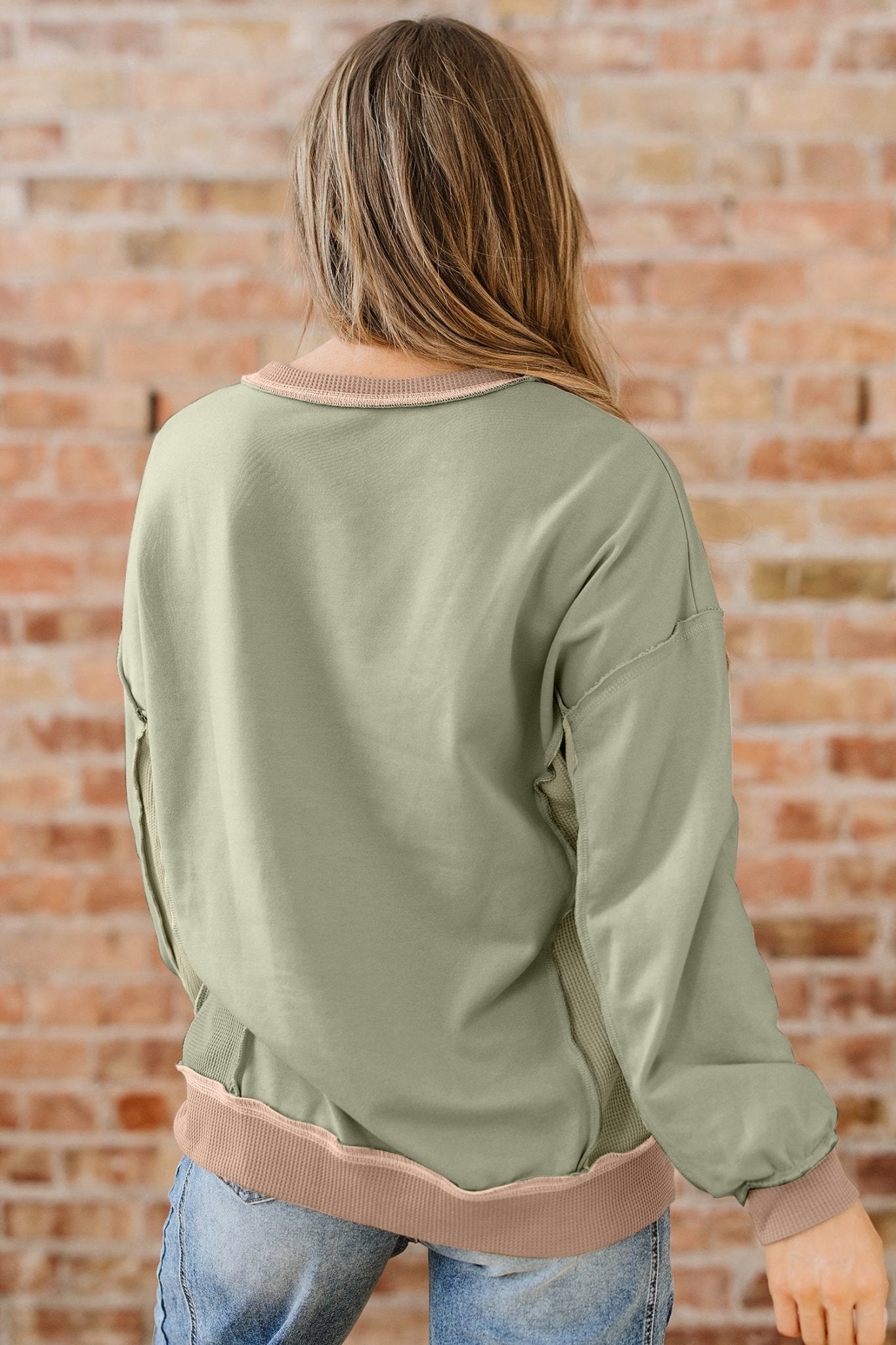Person wearing an olive green long-sleeve top with a brown waistband against a brick wall.
