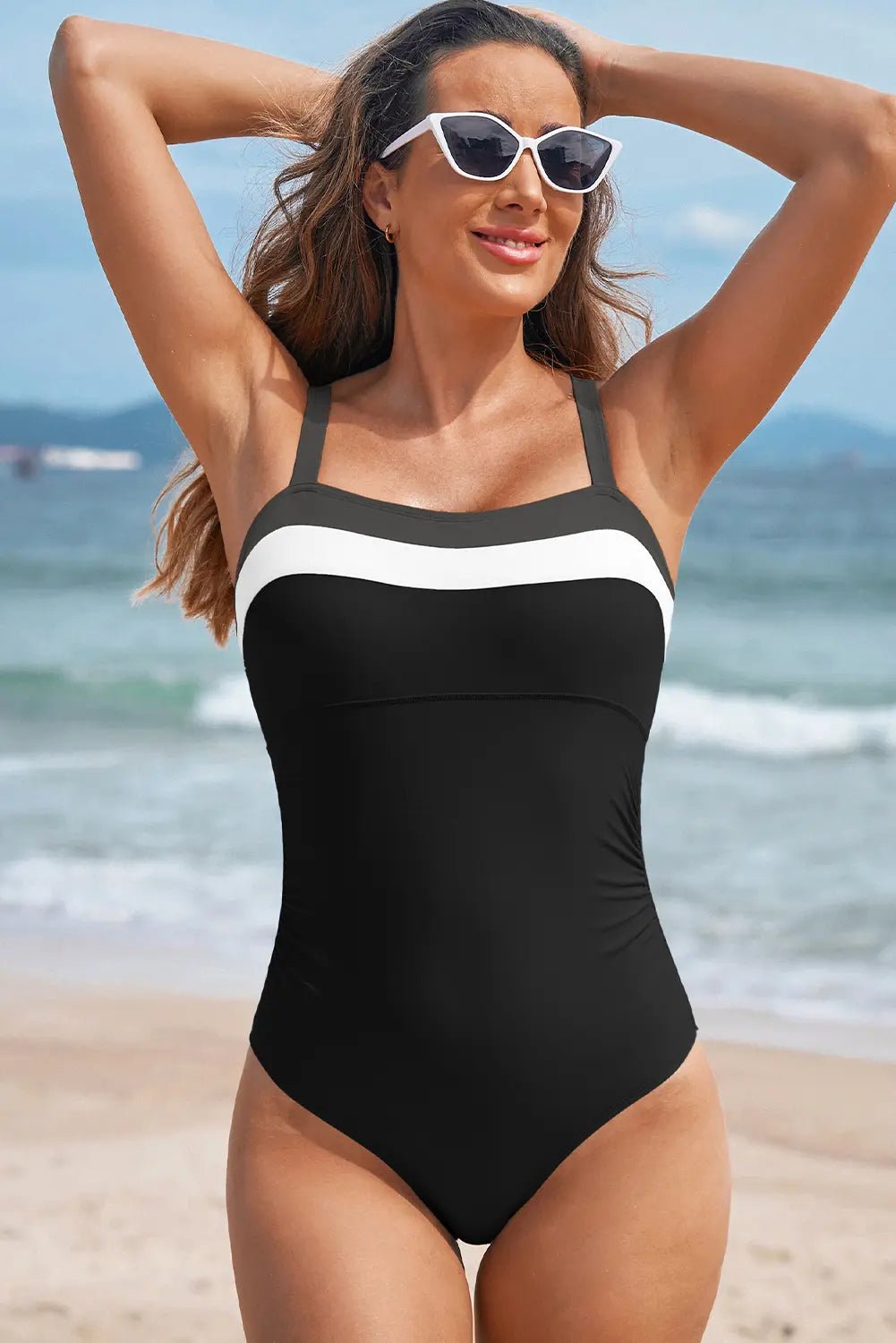 Woman wearing black one-piece swimsuit with white chest stripe and white sunglasses on a sandy beach.