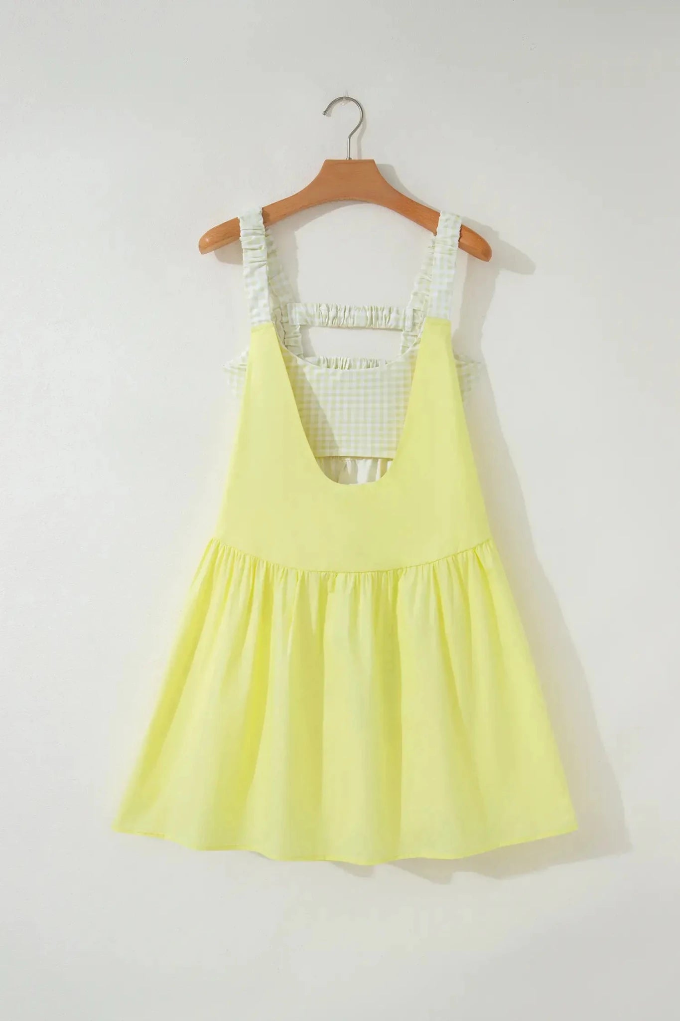 A light yellow sleeveless pinafore-style dress with a white and green gingham patterned inner bodice and shoulder straps, hanging on a wooden hanger against a plain off-white wall.