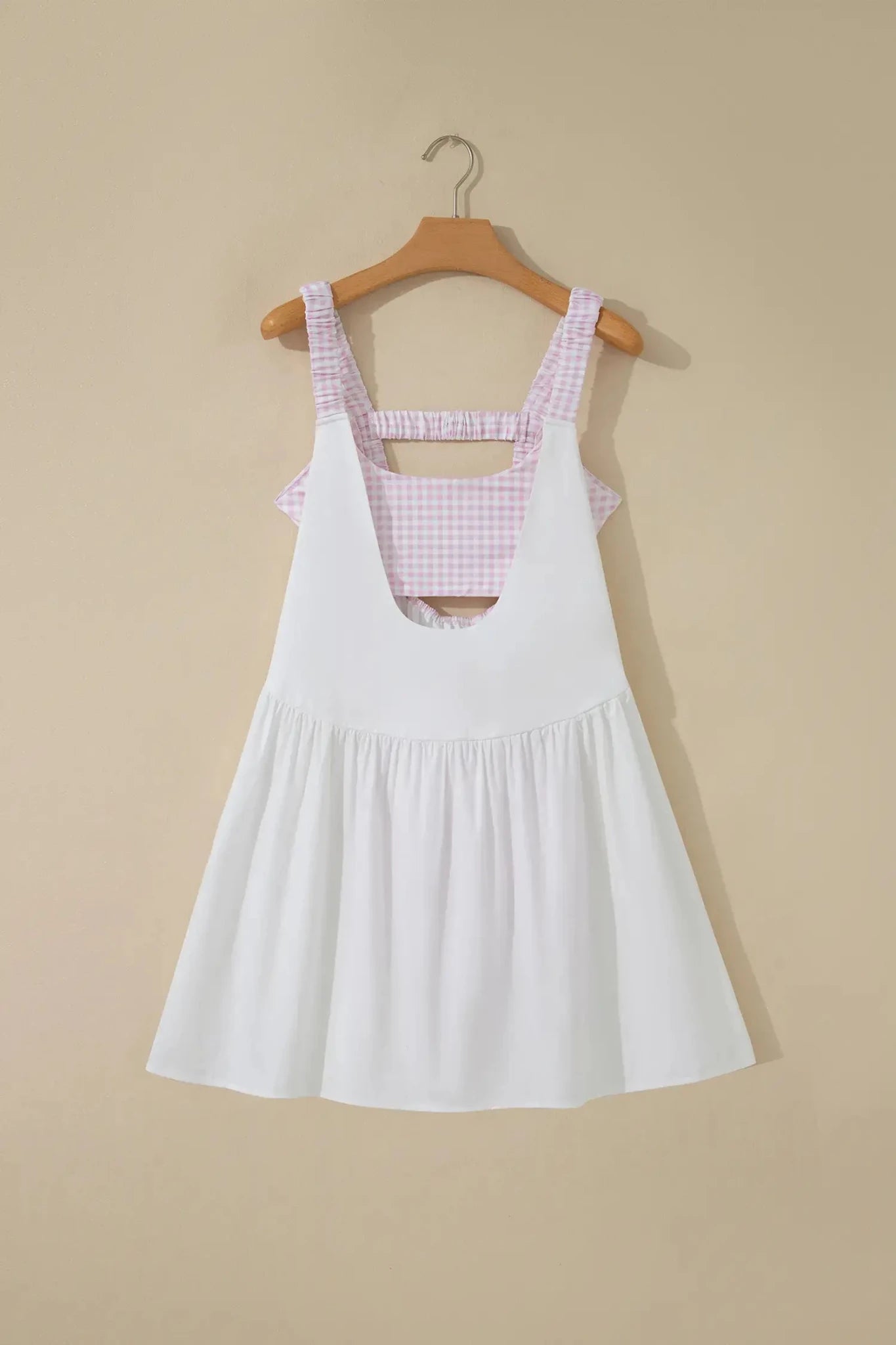 A white sleeveless pinafore-style dress with a pink and white gingham patterned underbust band and shoulder straps, hanging on a wooden hanger against a beige wall.