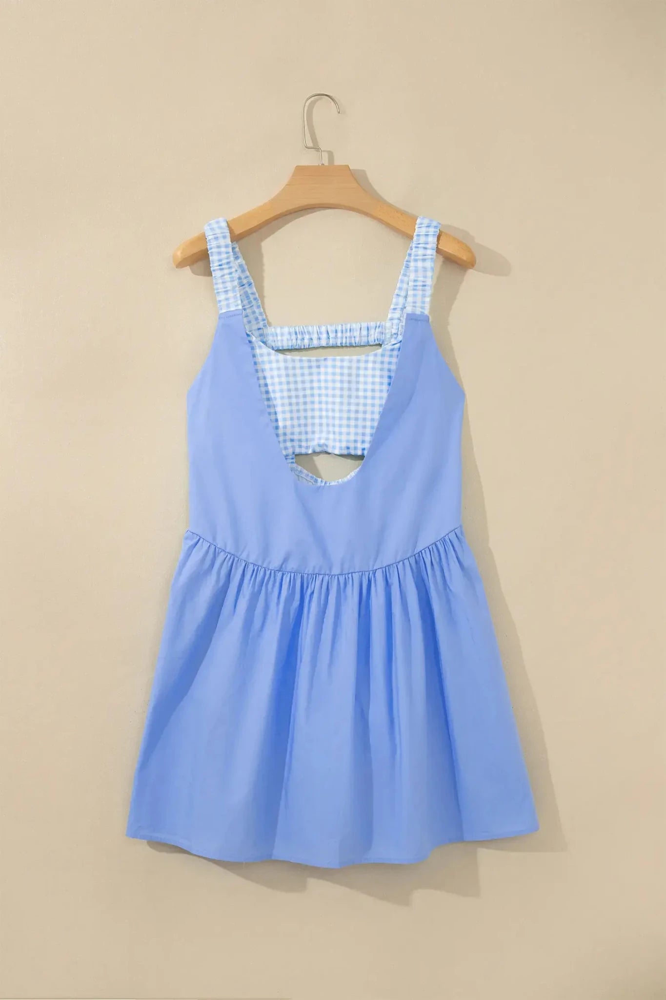 A light blue sleeveless dress with a blue and white gingham patterned neckline and straps, hanging on a wooden hanger against a beige wall.