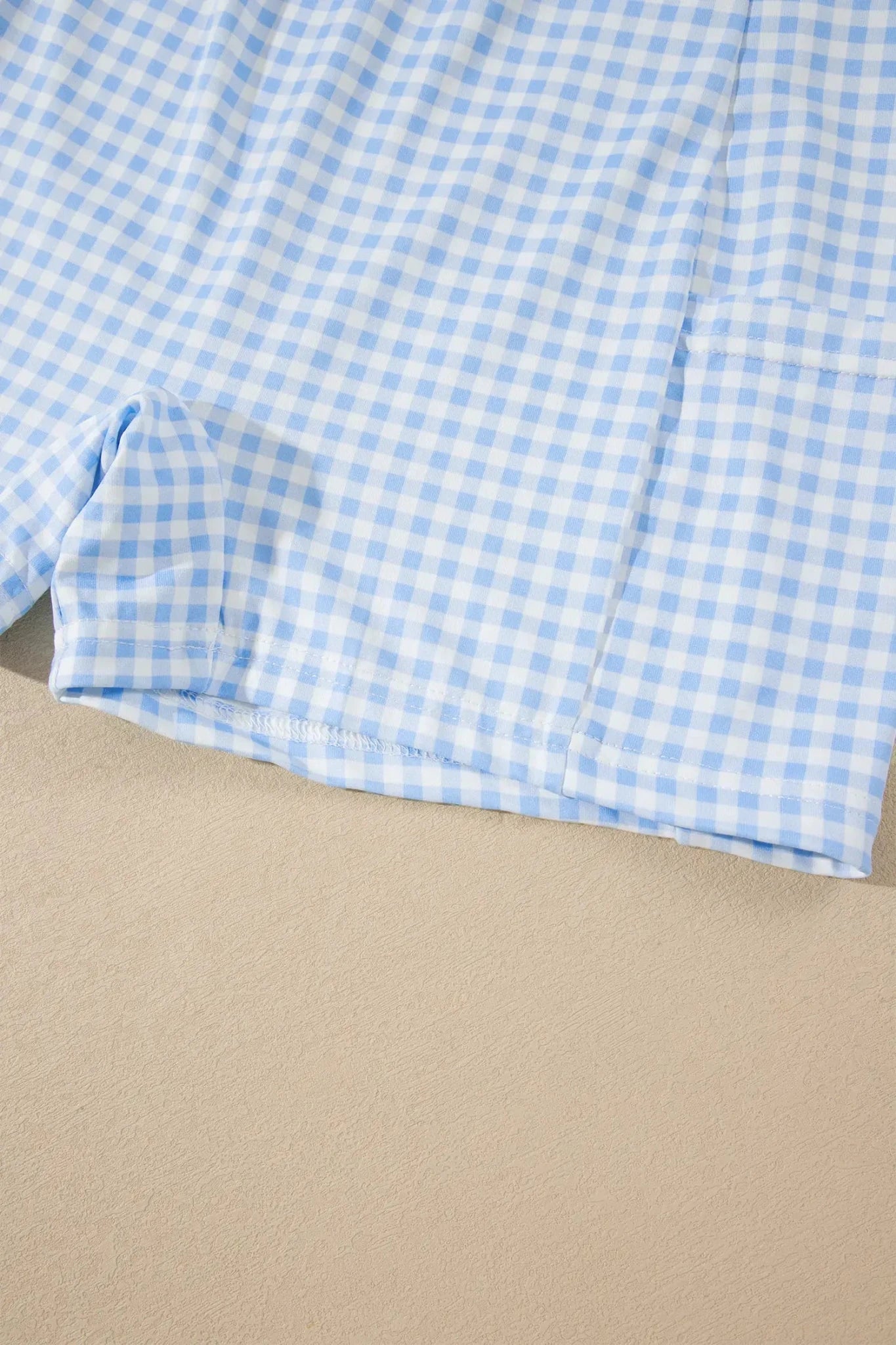 A close-up of a light blue and white gingham fabric, likely a shirt or shorts, laid flat on a beige surface.
