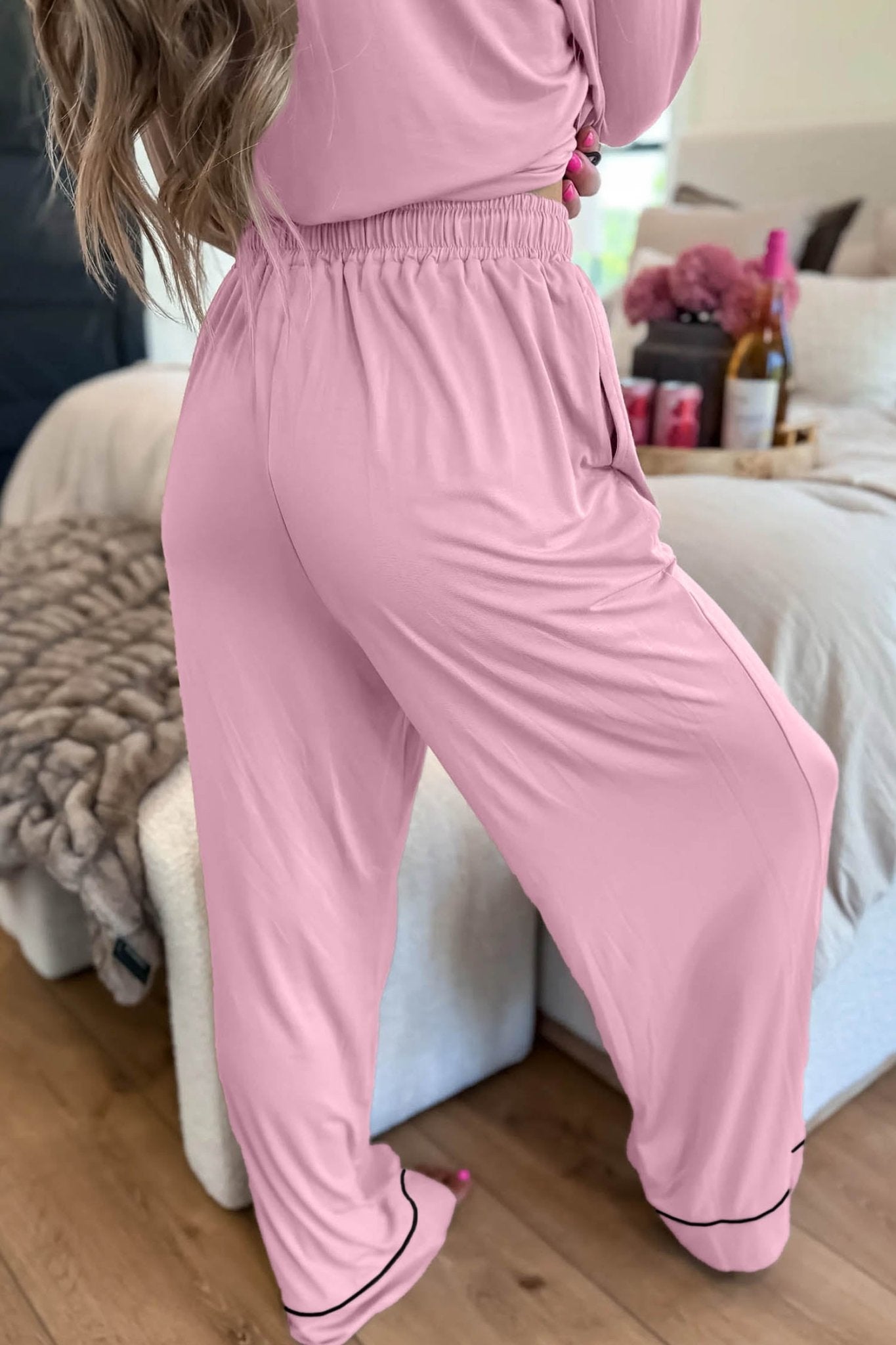 Woman in a pink pajama set with black piping, long-sleeve button-up top, and wide-leg pants in bedroom.