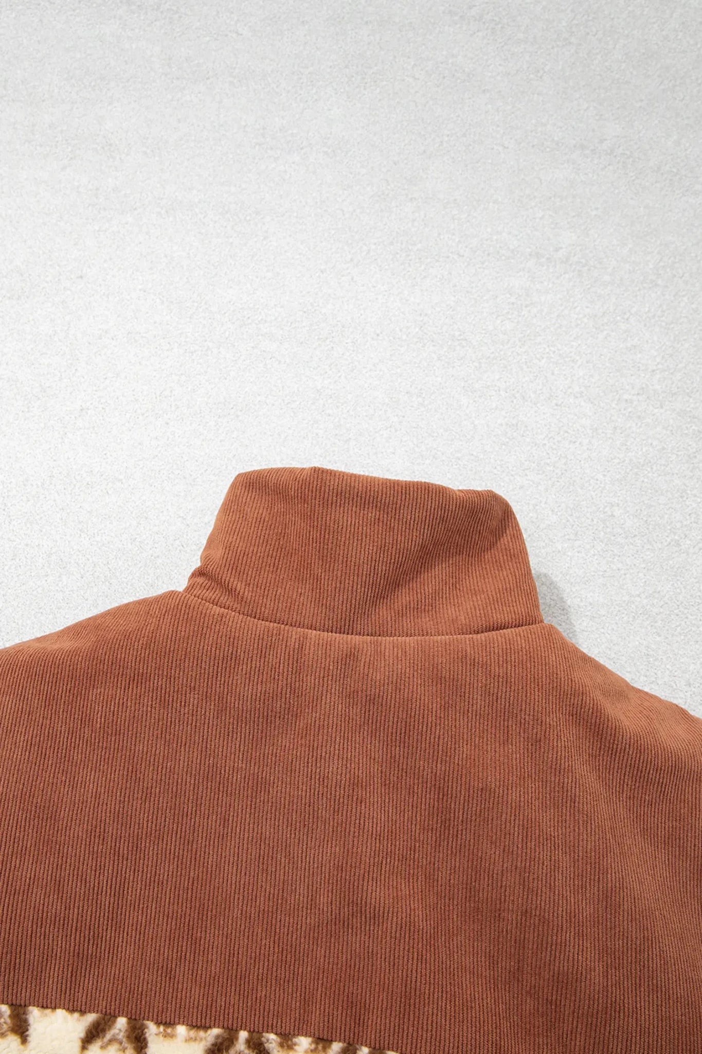 Close-up of the back collar of a rust-brown corduroy turtleneck sweater on a light gray textured background.