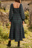 A woman wearing a long-sleeve plaid midi dress with a smocked bodice and tiered skirt, photographed from the back in an outdoor garden setting.