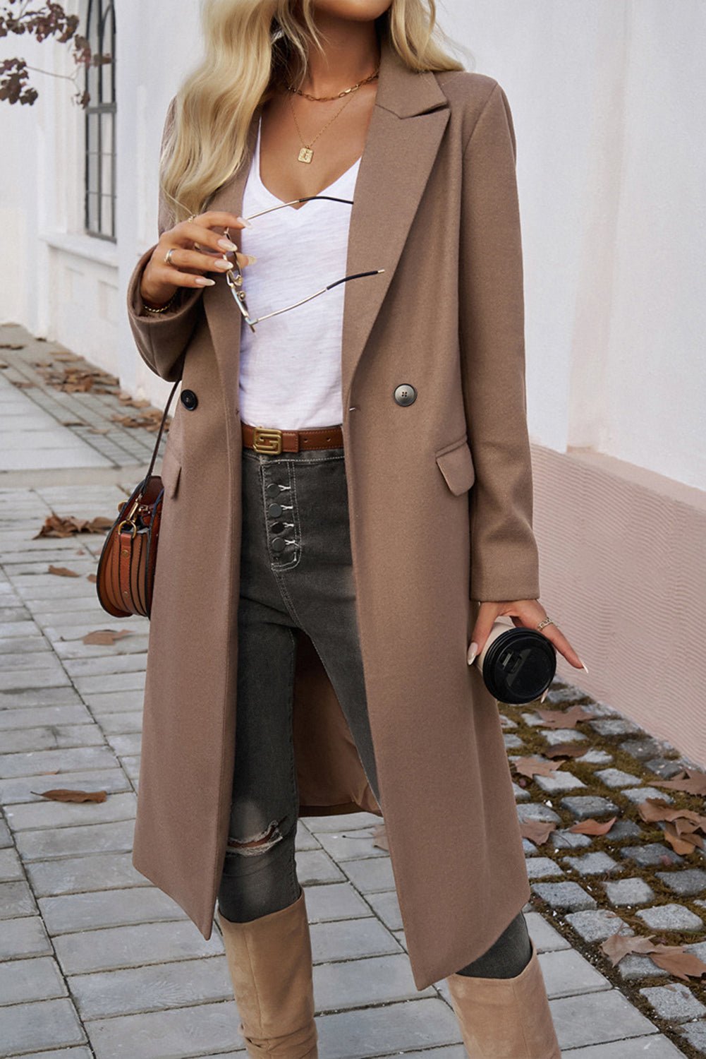 Woman wearing a long brown coat holding sunglasses and a coffee cup on a sidewalk.