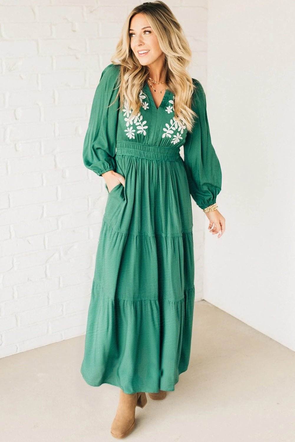 Woman wearing a green dress with floral embroidery against a white brick wall.