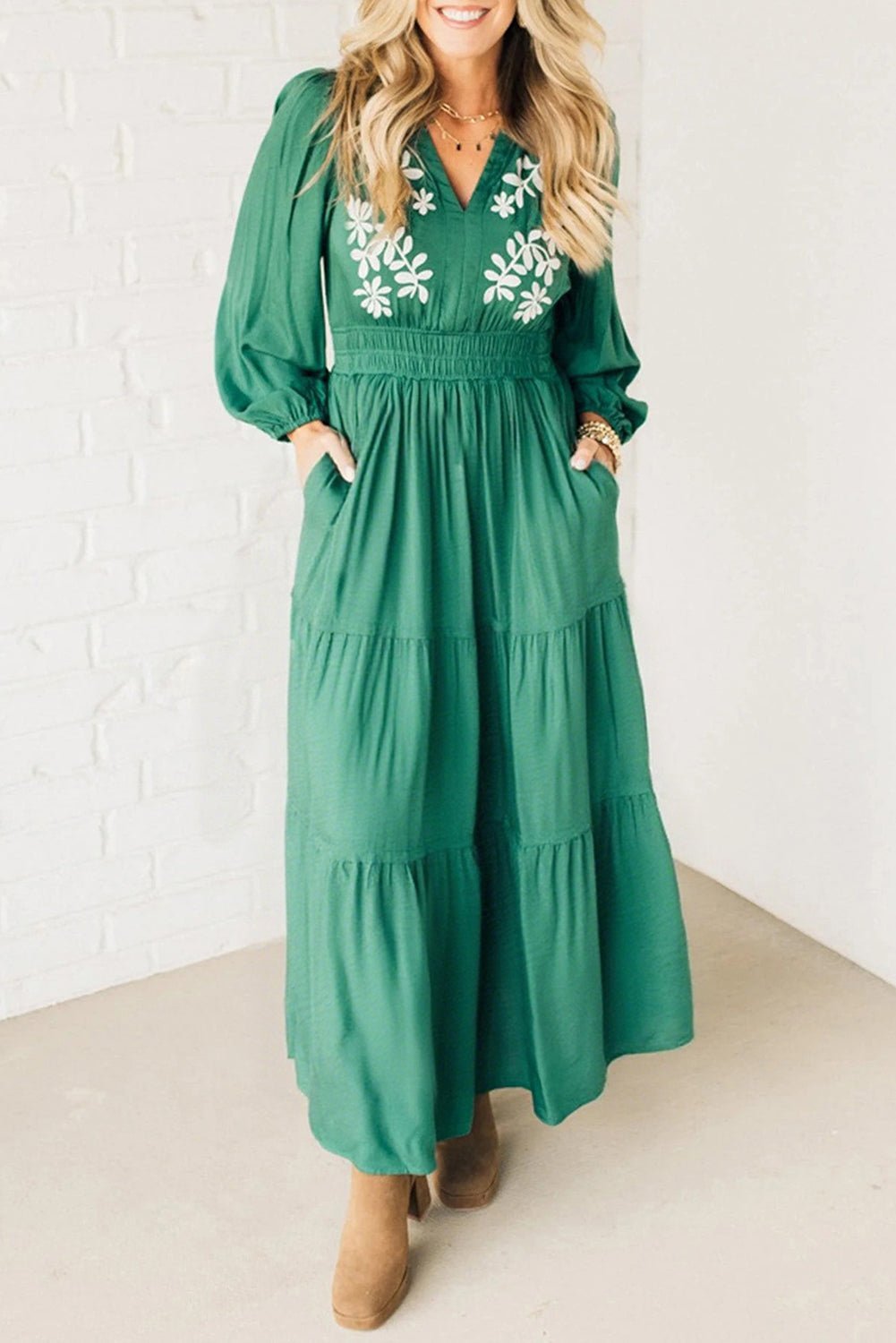 Woman wearing a green dress with floral embroidery against a white brick wall.