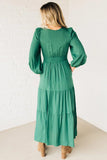 Woman wearing a green dress with long sleeves and a belt, standing against a white brick wall.