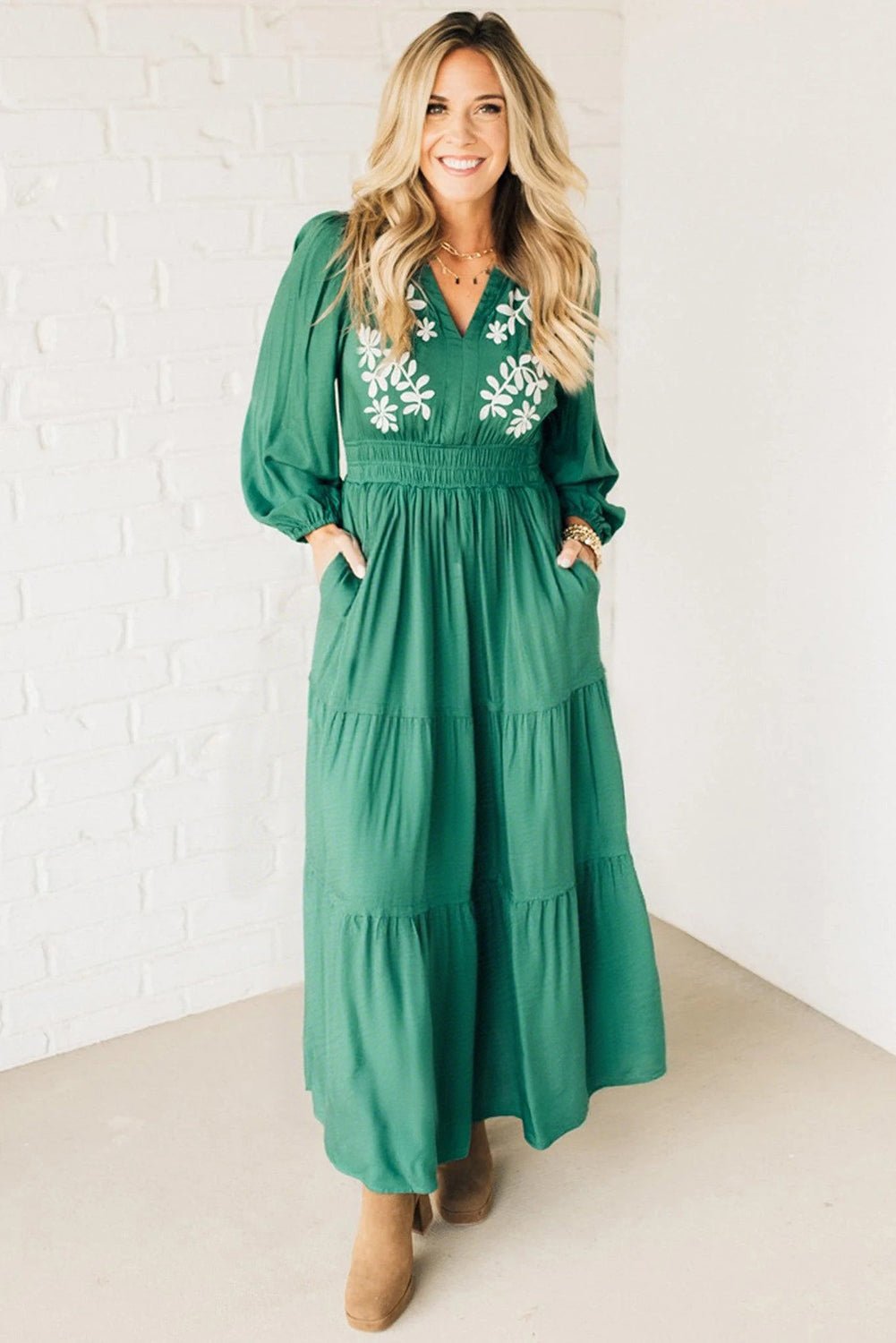 Woman wearing a green dress with floral embroidery against a white brick wall.