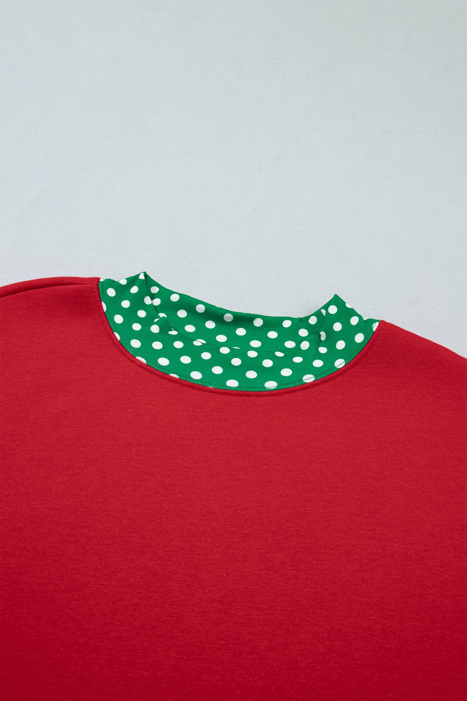 A close-up of a red crewneck top with a contrasting green collar featuring white polka dots, set against a light gray background.
