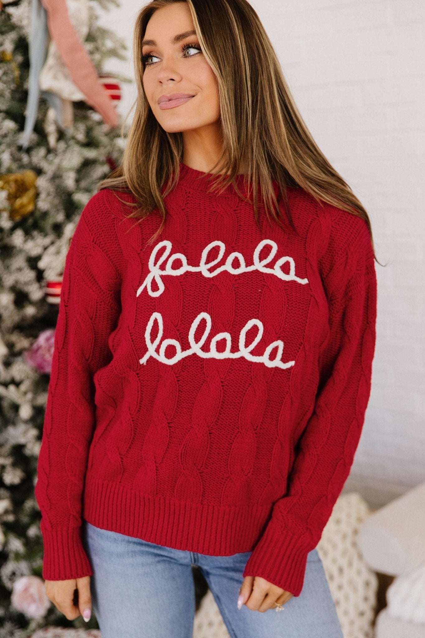 Woman in red cable-knit “fololo fololo” sweater with light blue jeans by Christmas tree.