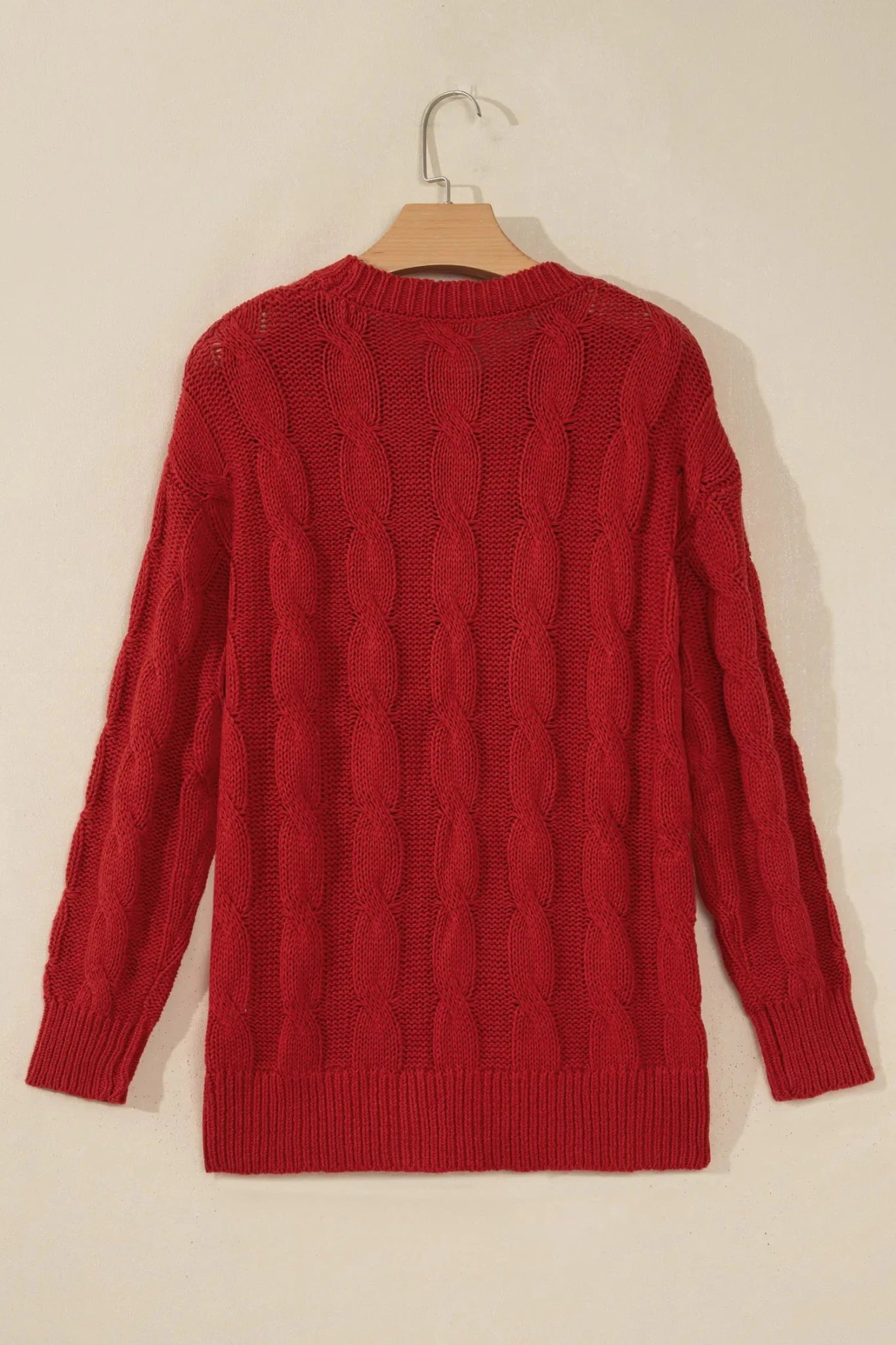 A red cable-knit sweater hanging on a wooden hanger against a plain beige wall.