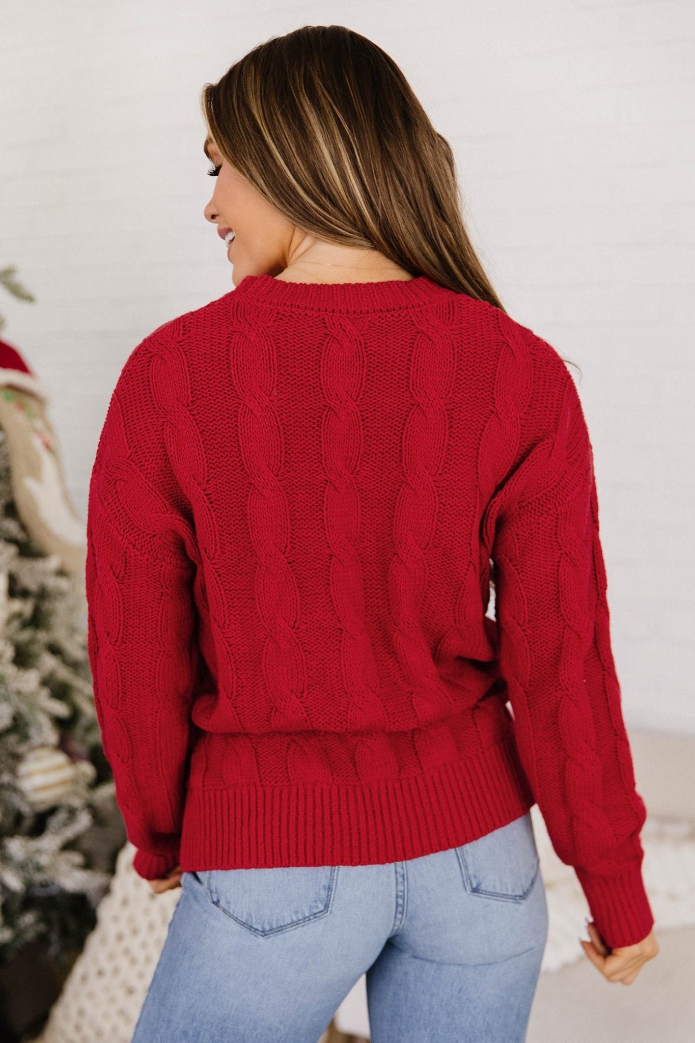 Back view of woman in red cable-knit sweater and light blue jeans by Christmas tree.