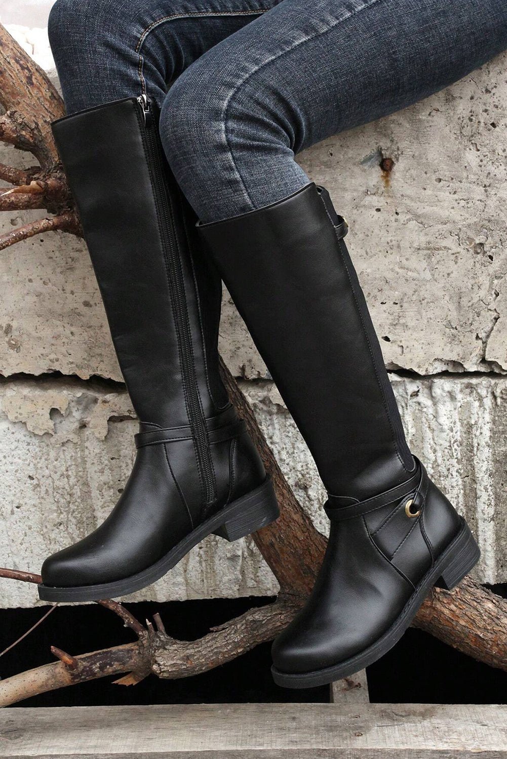 Person wearing black knee-high boots sitting on a branch with a concrete wall background
