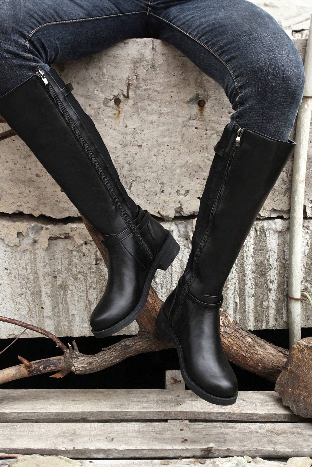 Black knee-high boots worn with blue jeans against a rustic stone wall.