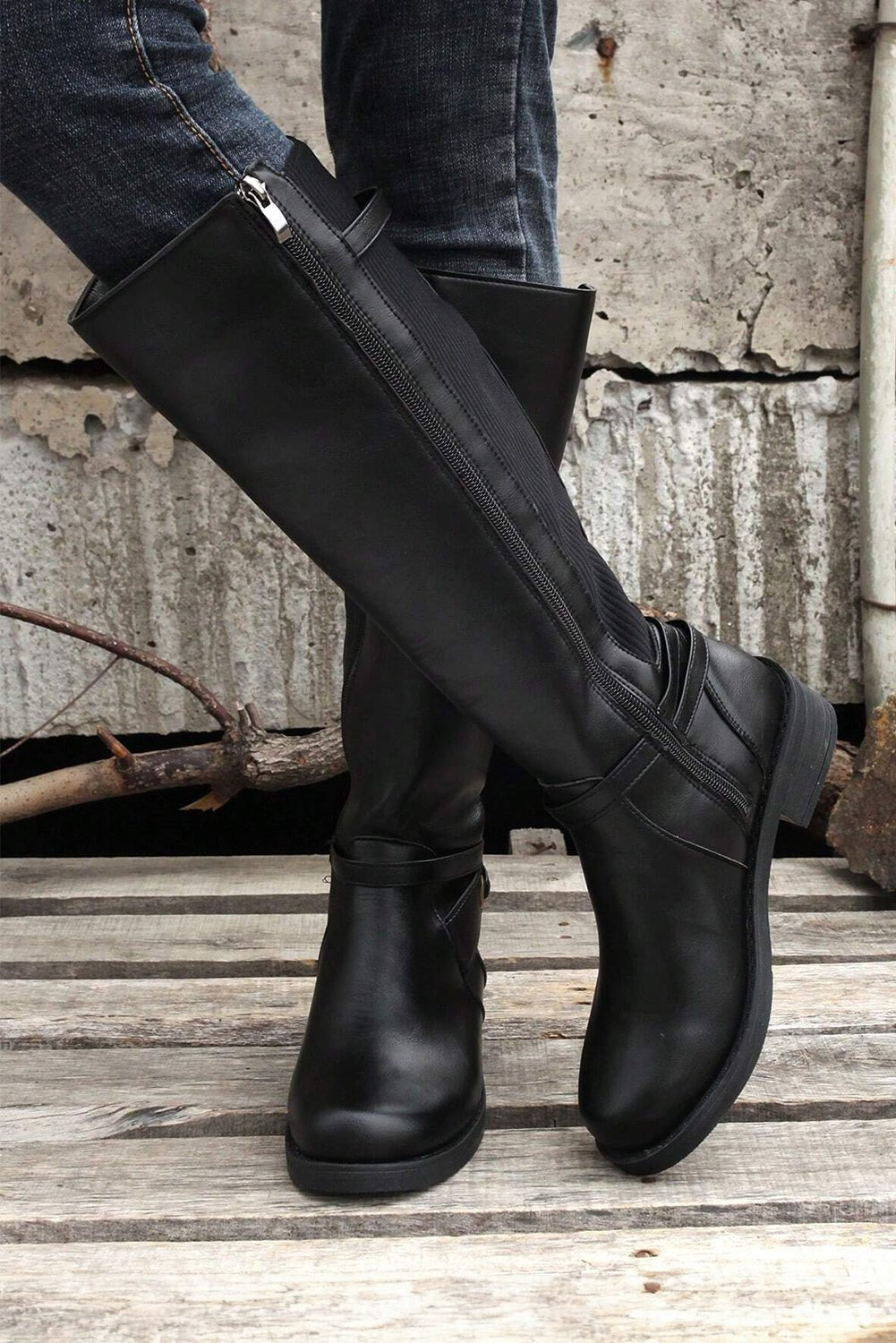 Person wearing black knee-high boots standing on a wooden floor with a concrete wall in the background.