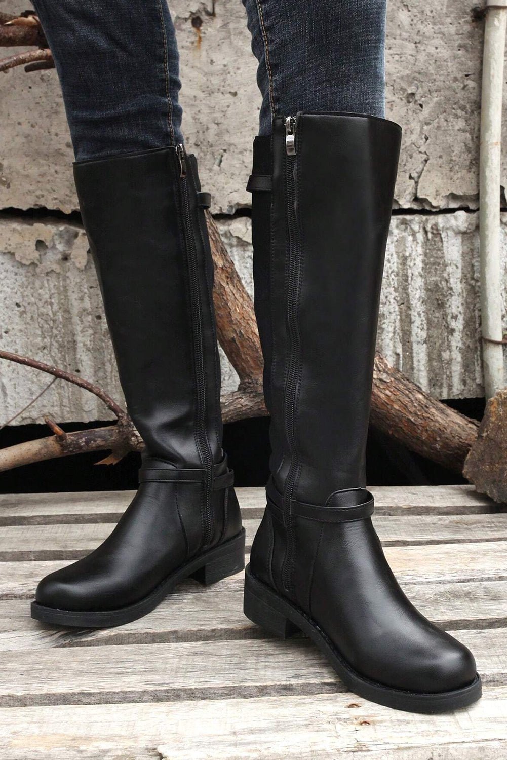 Black knee-high boots worn by a person standing on a wooden floor with a rustic background.
