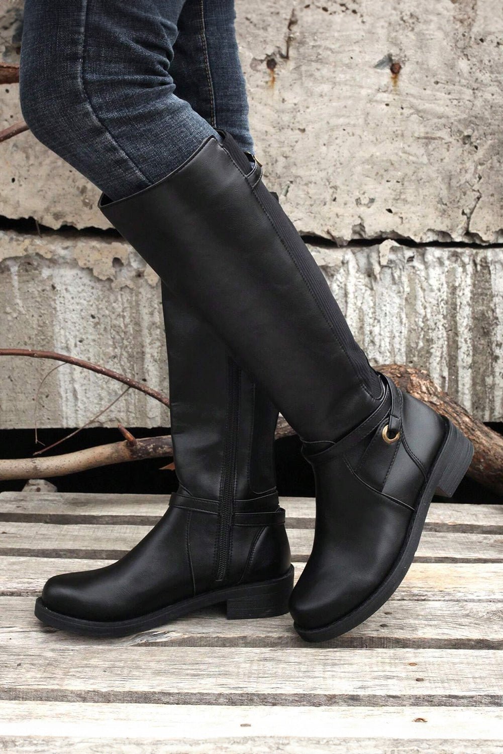 Person wearing black knee-high boots standing on a wooden surface with a concrete wall in the background.