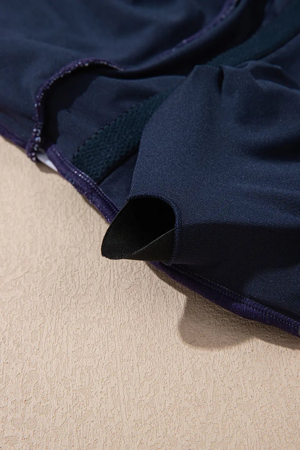Close-up of dark navy blue garment showing fabric texture, mesh panel, and dark piping on a textured beige surface.