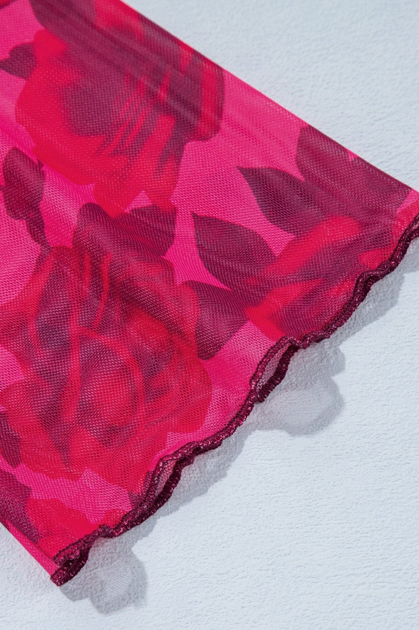 Close-up of sheer mesh fabric with dark red rose pattern on bright pink with scalloped edge on light gray.