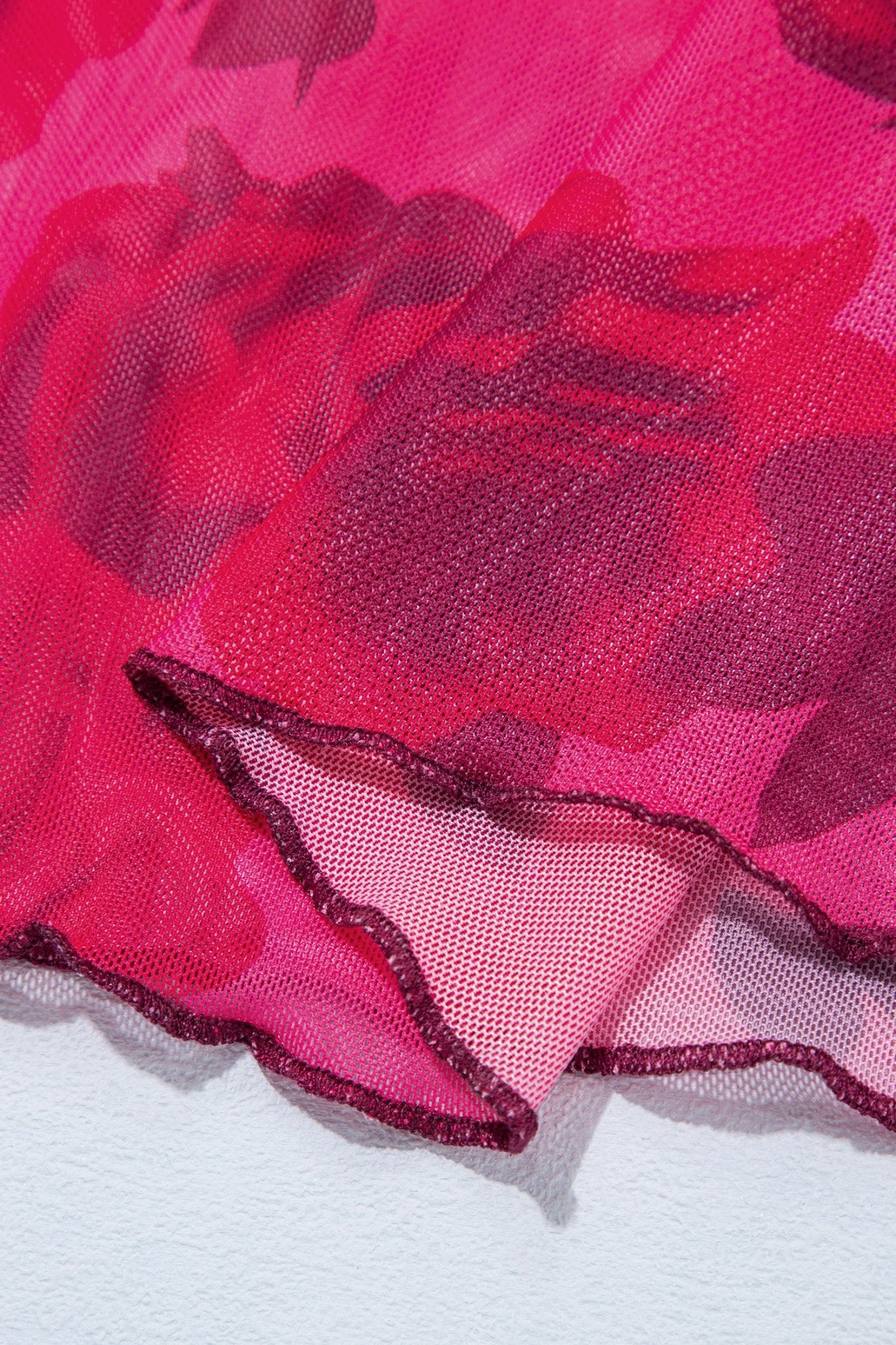 A close-up of a sheer pink mesh fabric with a darker pink floral pattern and a ruffled edge, laid over a white surface.