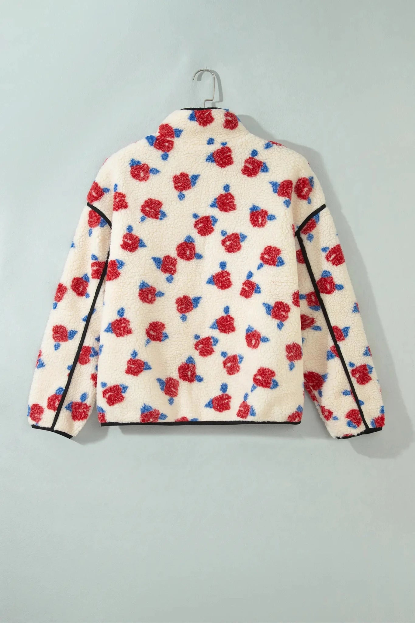 A cream-colored fleece jacket with a red and blue rose pattern, hanging on a metal hanger against a light gray wall.