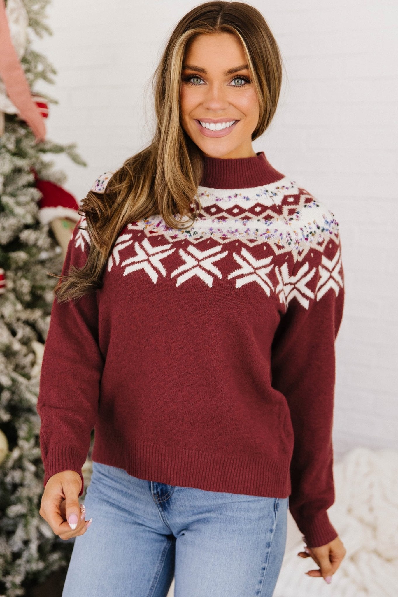 Woman wearing a burgundy Fair Isle Christmas sweater with blue jeans by a decorated tree.