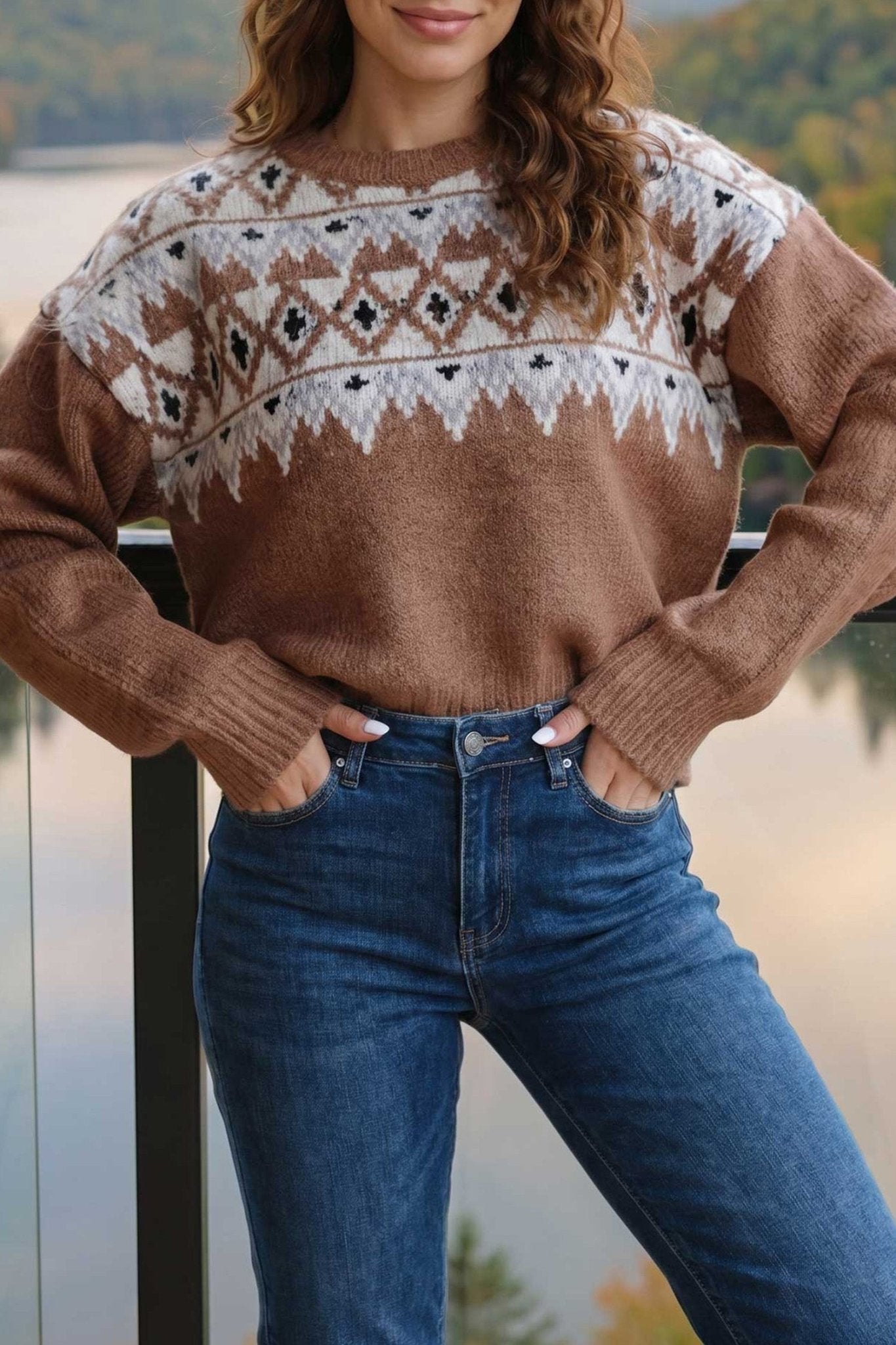 Woman in brown Fair Isle knit sweater with cream-black pattern and blue jeans by a lake.