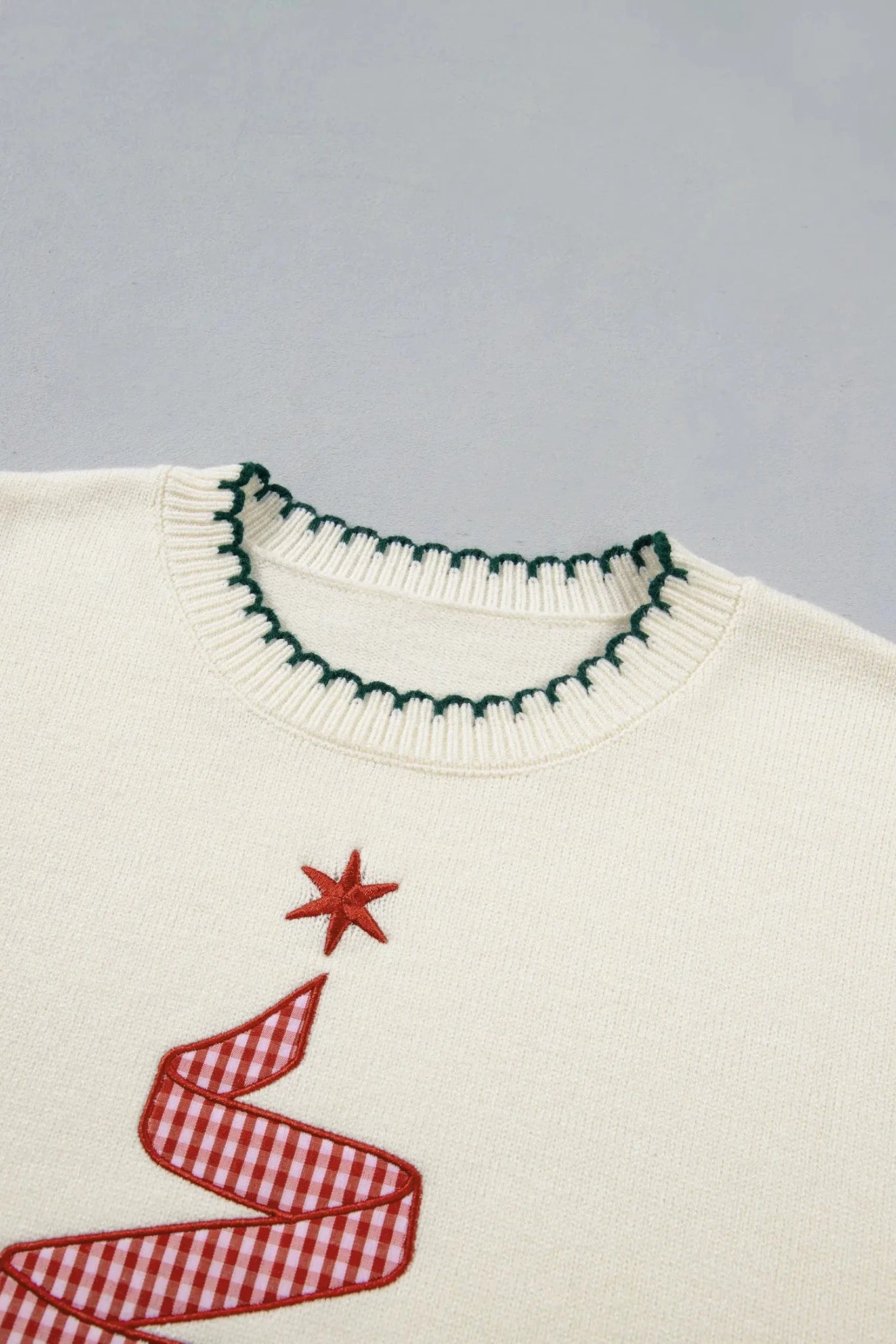Close-up of an off-white knit sweater with a green scalloped trim at the collar and a red-and-white gingham Christmas tree motif with a red star on the chest, set against a light gray background.