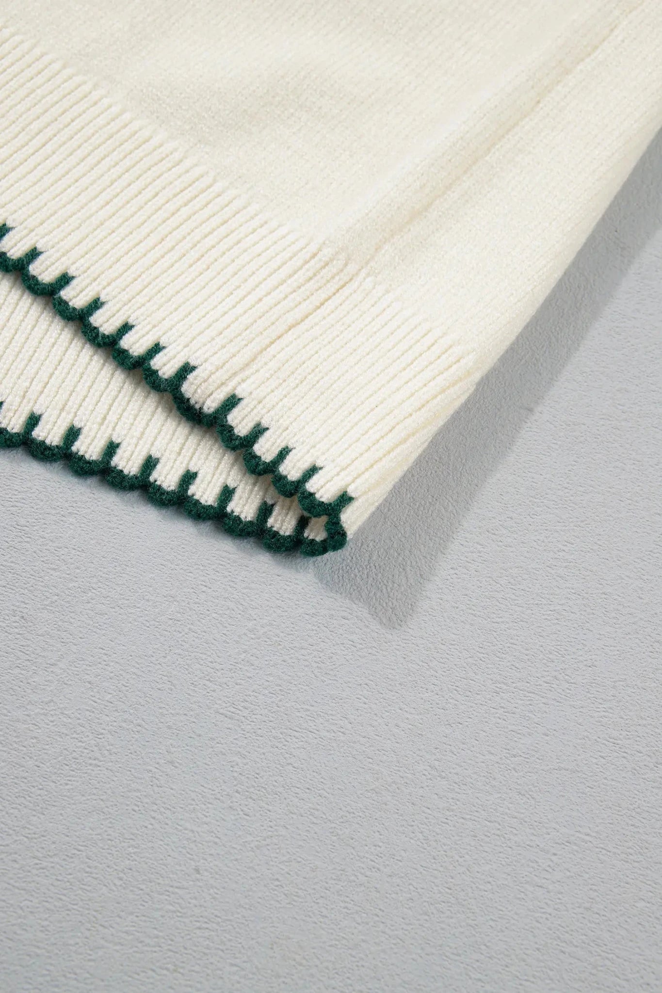 Close-up of a cream-colored ribbed knit fabric with a dark green zigzag stitched border, laid flat on a light gray textured surface.