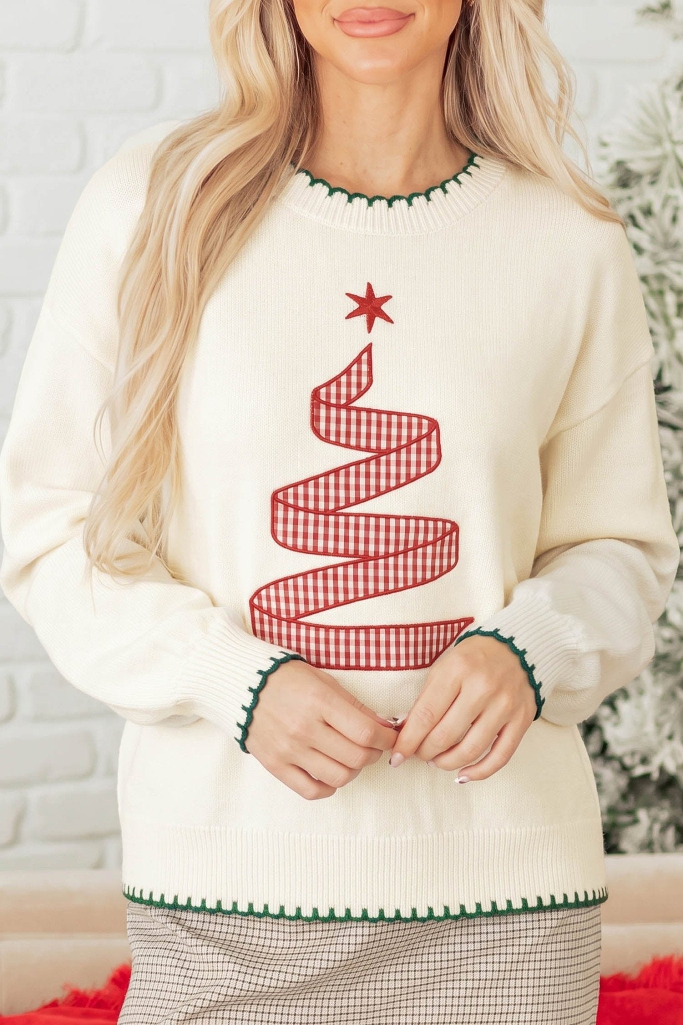 A woman wearing a cream-colored crewneck sweater with a red and white gingham Christmas tree design on the front, featuring green trim at the collar and cuffs.