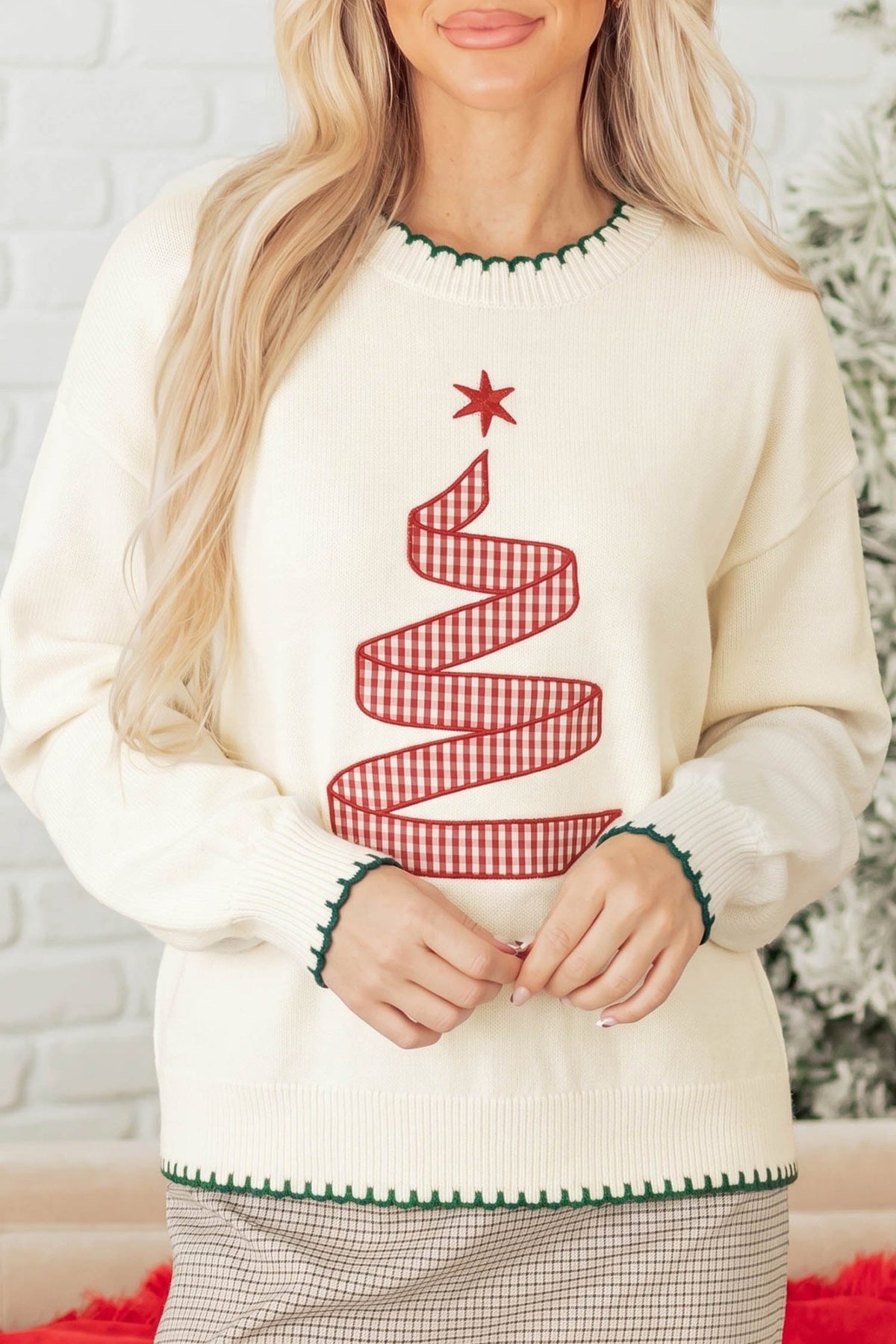 A woman wearing a cream-colored crewneck sweater with a red and white gingham Christmas tree design on the front, featuring green trim at the collar and cuffs.