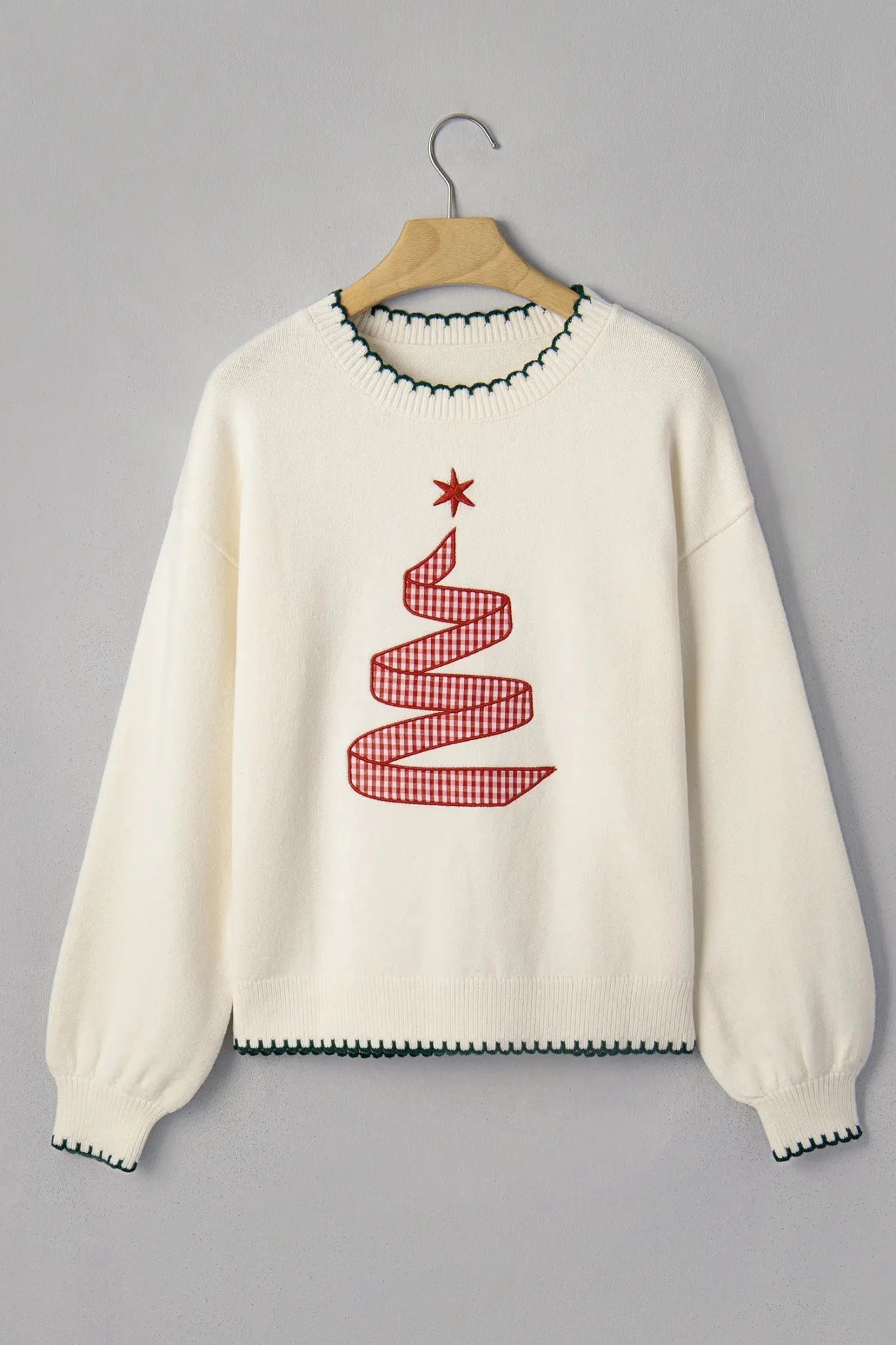 An off-white knit crewneck sweater hanging on a wooden hanger against a solid gray background. The sweater features a red and white gingham Christmas tree motif with a red star on the chest, and dark green contrast knit trim along the collar, hem, and cuffs.