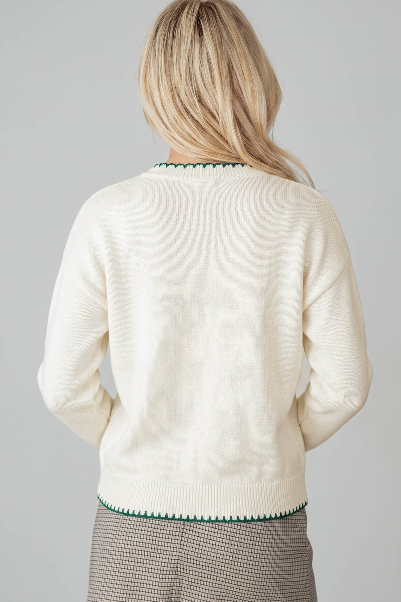 A woman viewed from the back wearing an off-white knit sweater with green trim at the collar and hem, paired with a brown and white checkered skirt, against a solid gray background.
