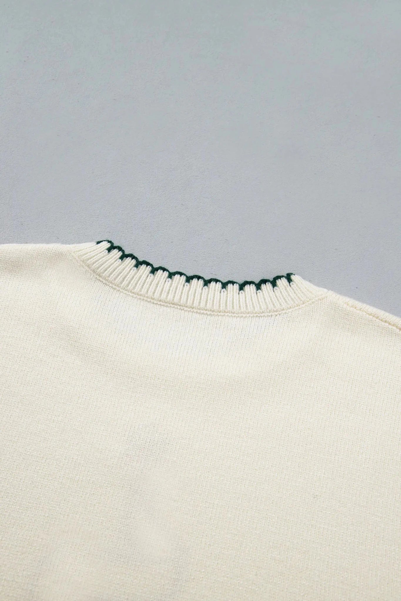 Close-up of a cream-colored knit sweater showing the ribbed crew neck with a distinctive dark green zigzag stitch detail along the collar edge, set against a solid light gray background.