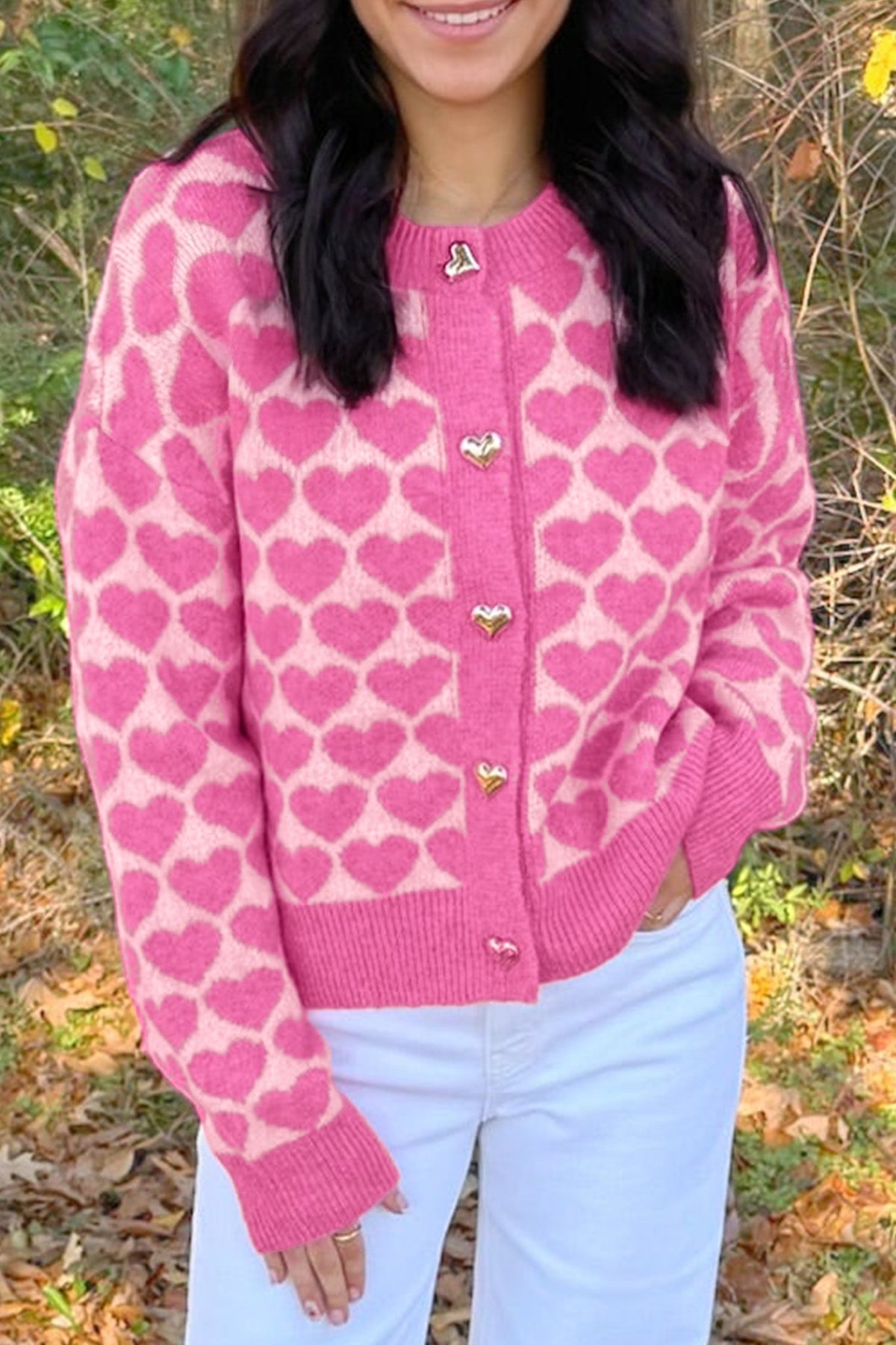 Woman in pink heart-patterned cardigan with gold heart buttons and white pants.

