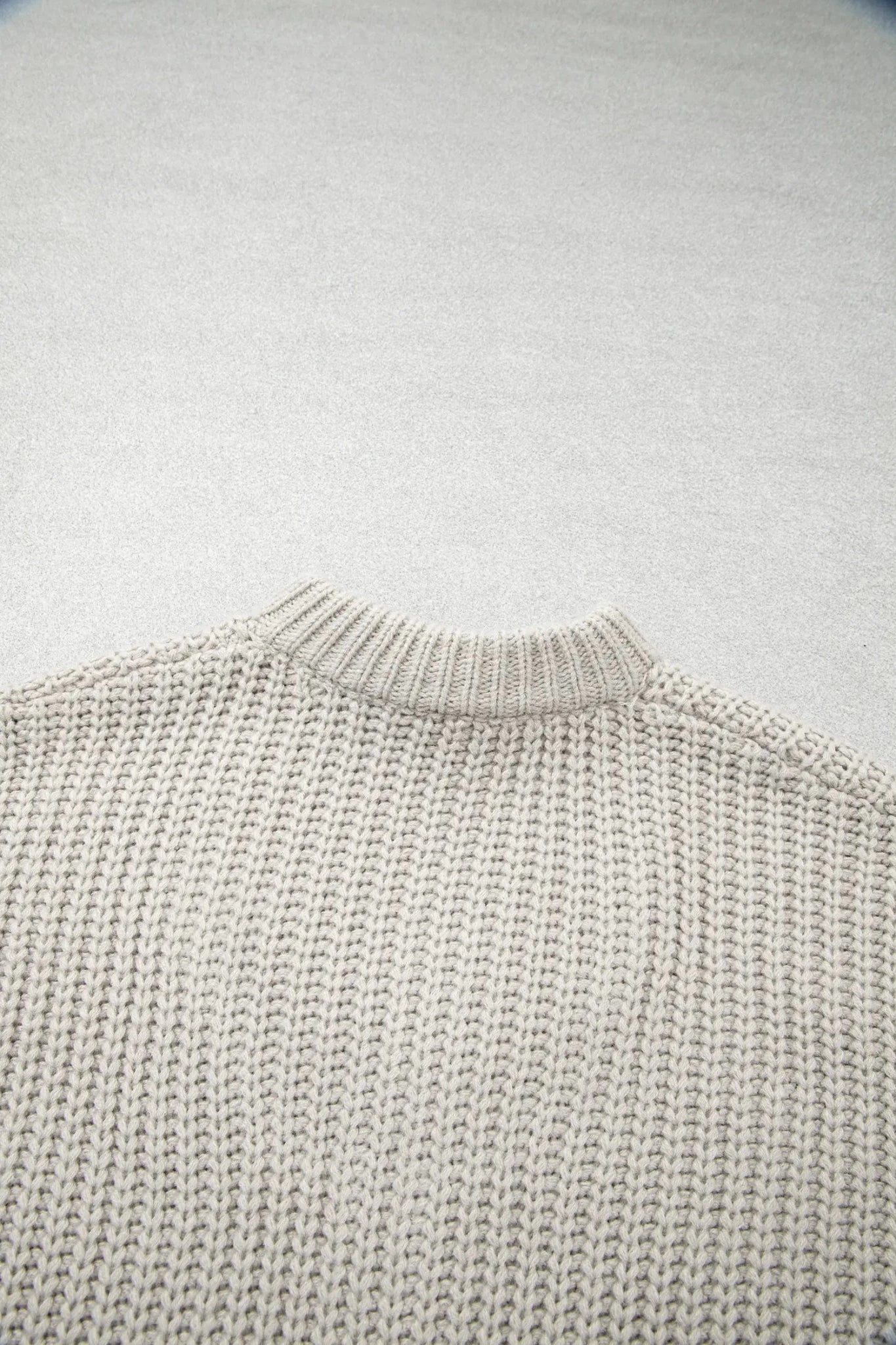 Close-up of a light oatmeal knit sweater with a ribbed crew neck on gray.
