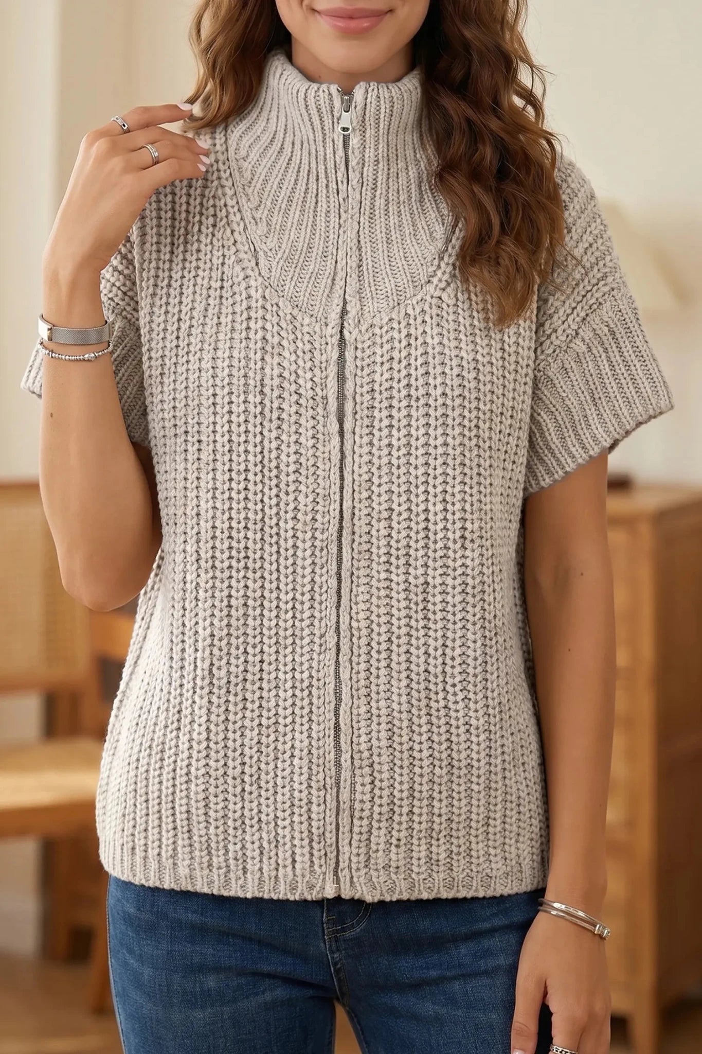 A woman wearing a chunky knit short-sleeve zip-front vest in a light heather gray color, paired with blue jeans.
