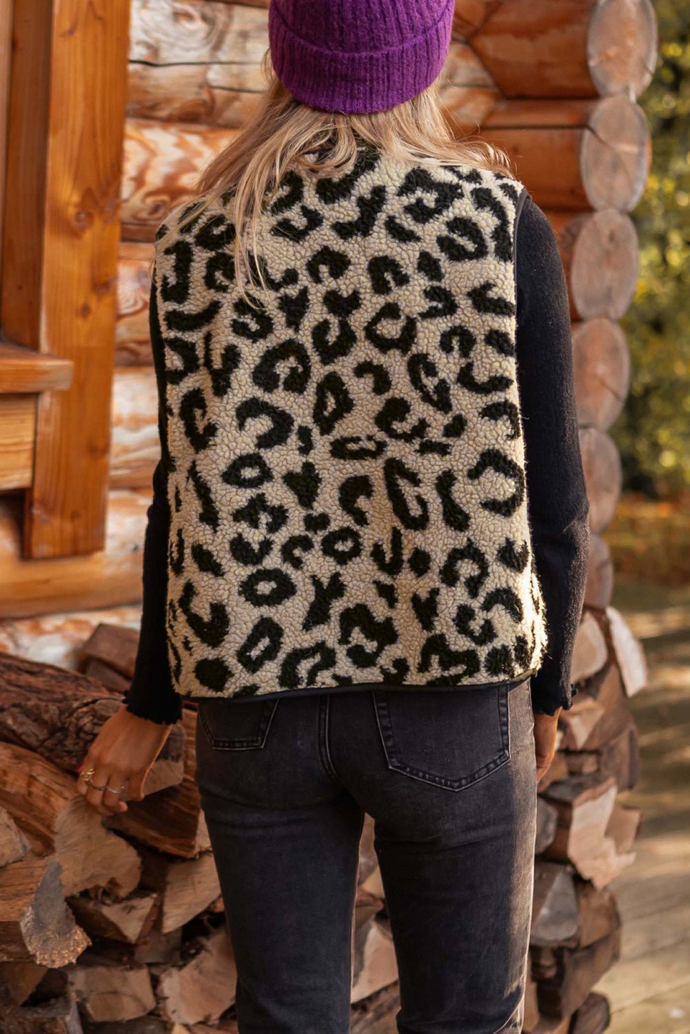 Person wearing a leopard print vest and purple beanie standing in front of stacked logs.