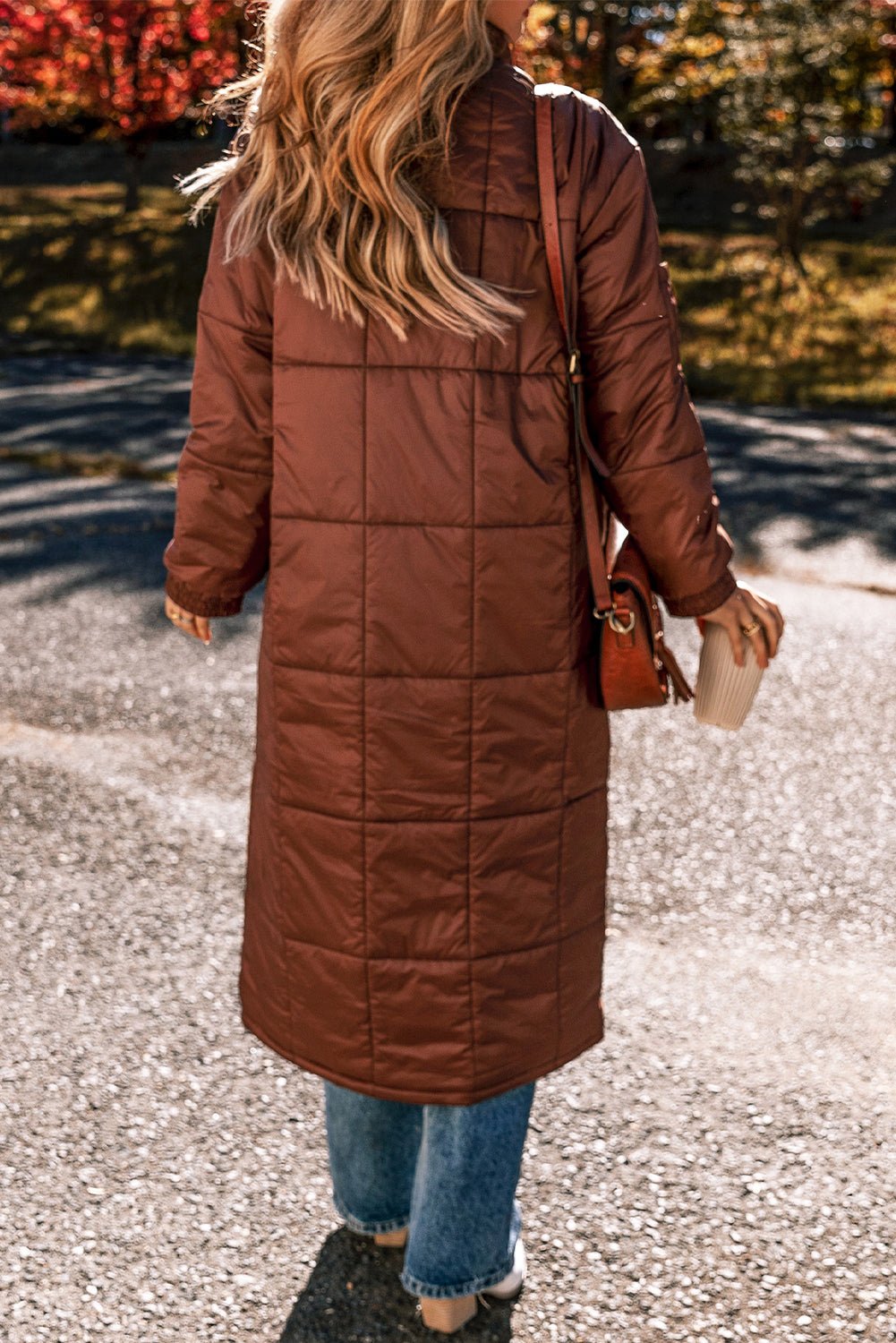 Person wearing a brown quilted coat walking outdoors with autumn foliage in the background