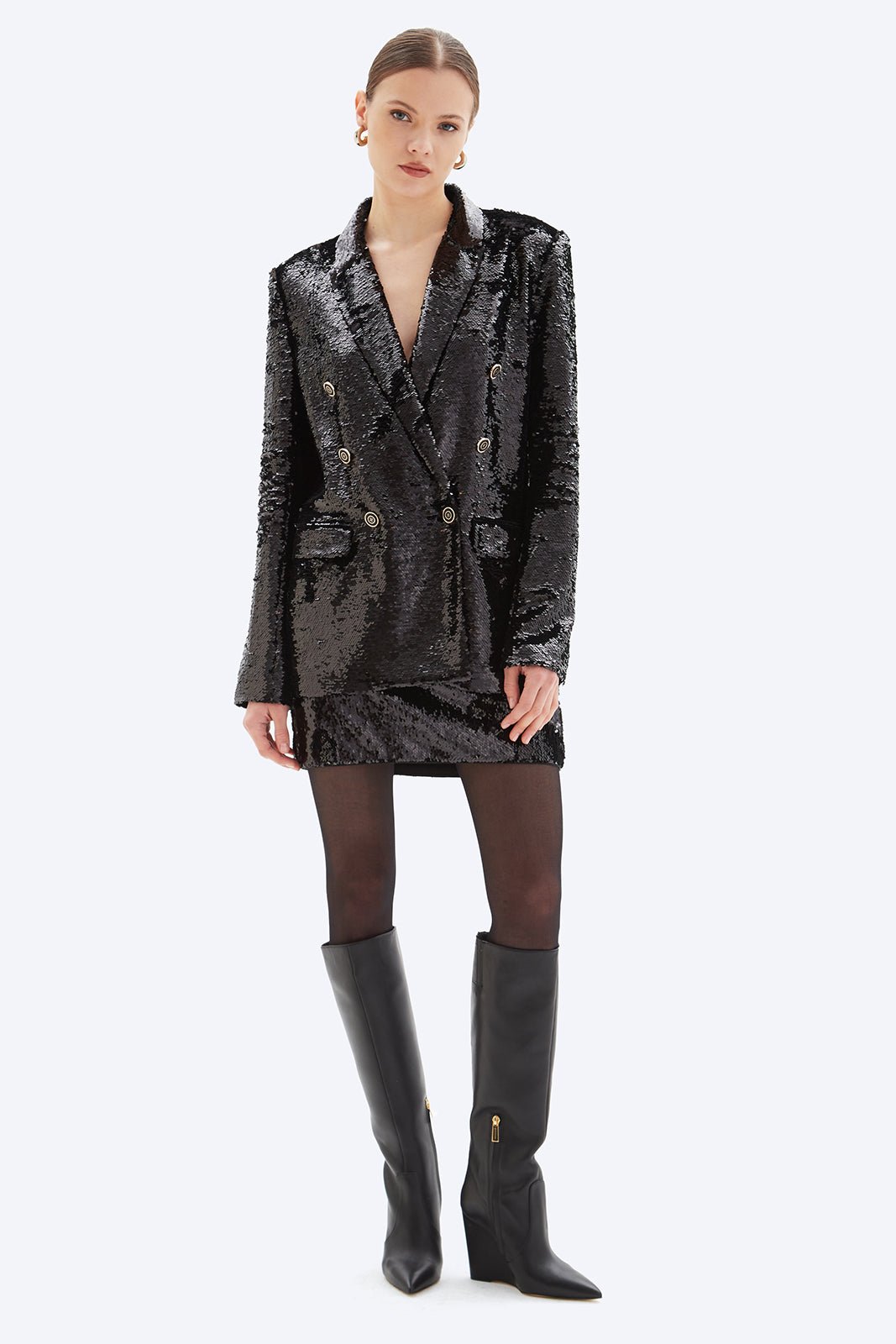 Woman wearing a black sequin blazer dress with knee-high black boots on a white background