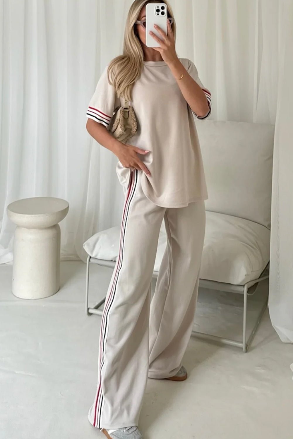 Woman taking a mirror selfie wearing a beige outfit with white pants and red stripes in a minimalistic room.
