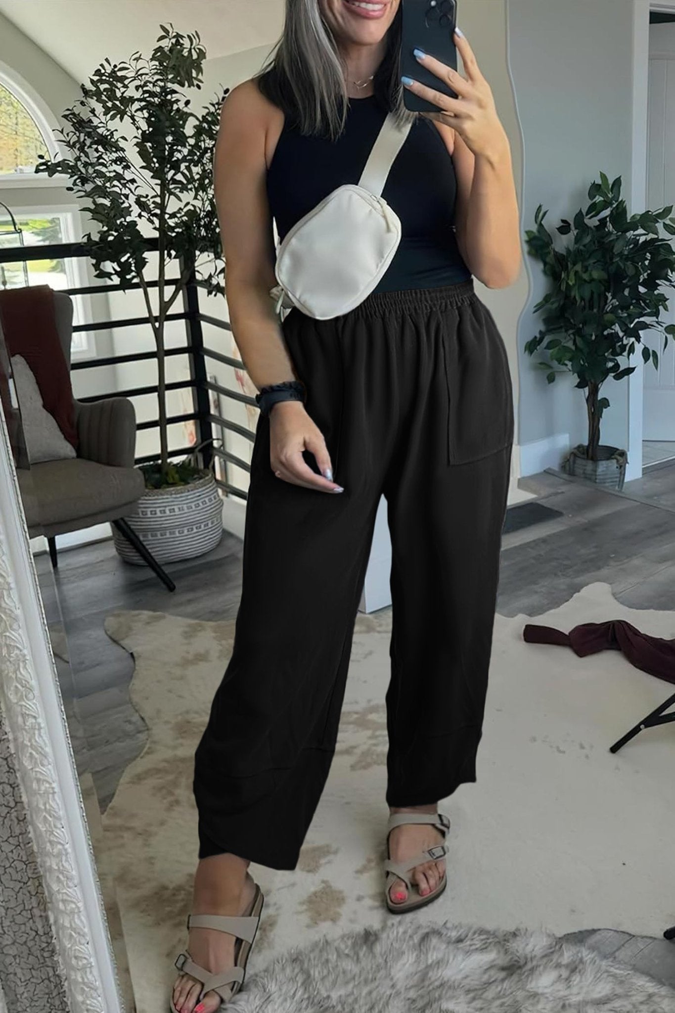 Person taking a mirror selfie wearing a black sleeveless top and wide-leg pants in a room with plants and furniture.