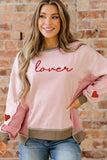 Woman wearing a pink sweatshirt with 'lover' text and heart designs, standing against a brick wall.