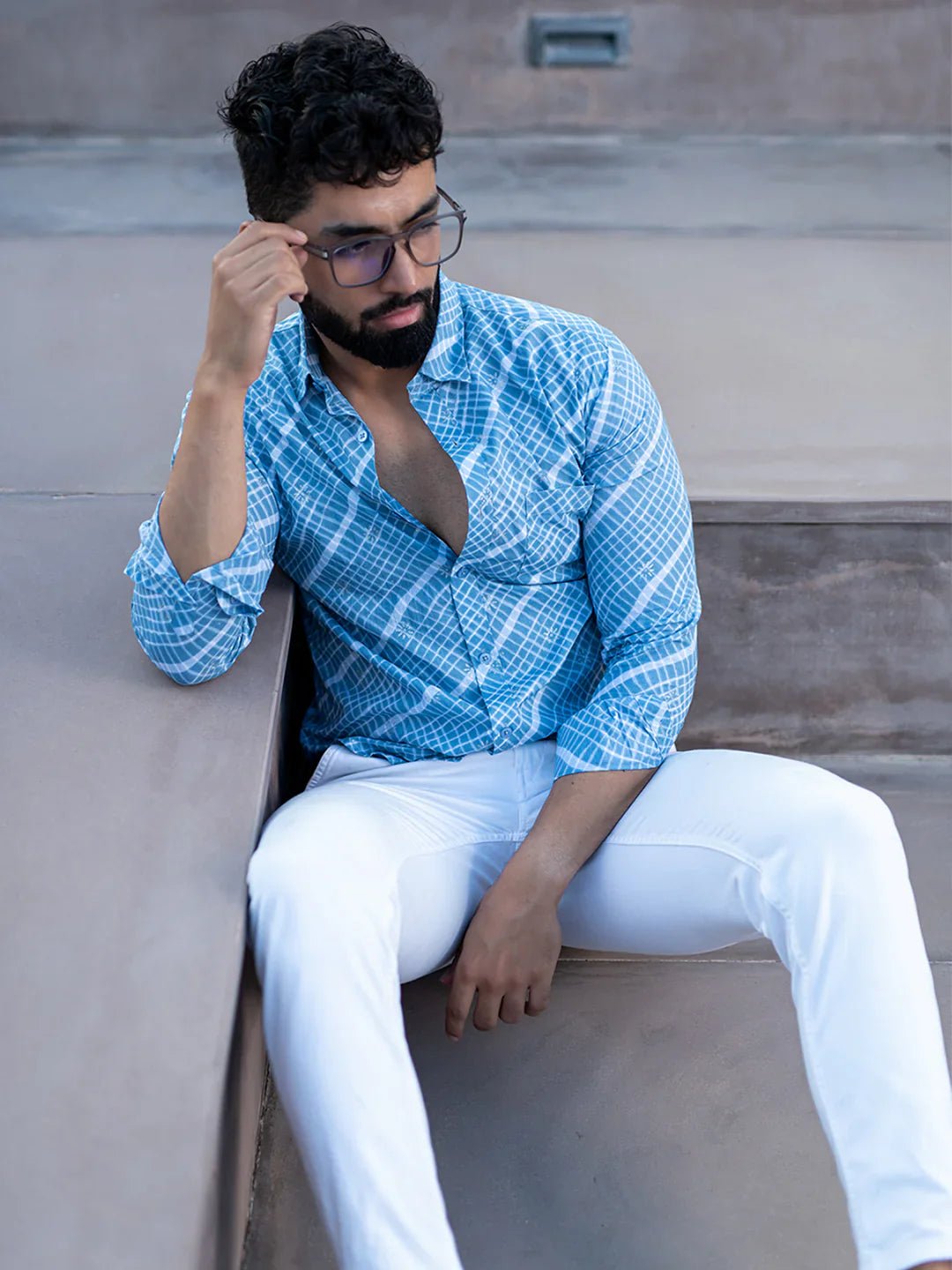 Man wearing a blue patterned shirt and white pants sitting on a bench.