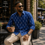 Man in a blue patterned shirt sitting outdoors holding a coffee cup.