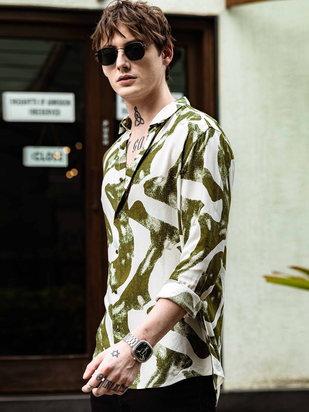 Person wearing a green and white patterned shirt with sunglasses and a tattoo.