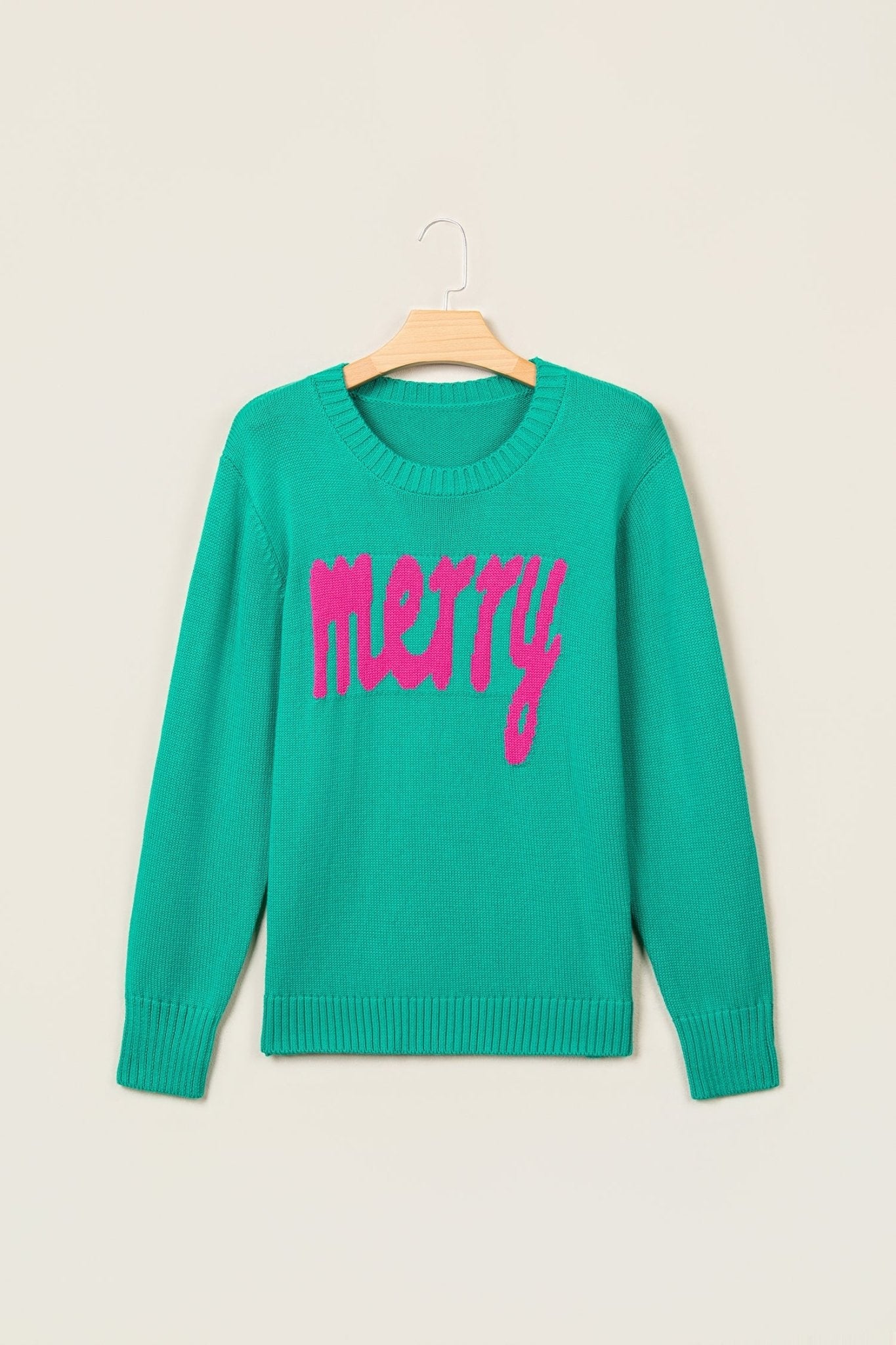 Teal green crewneck sweater with “merry” in bright pink script, hanging on a wooden hanger.